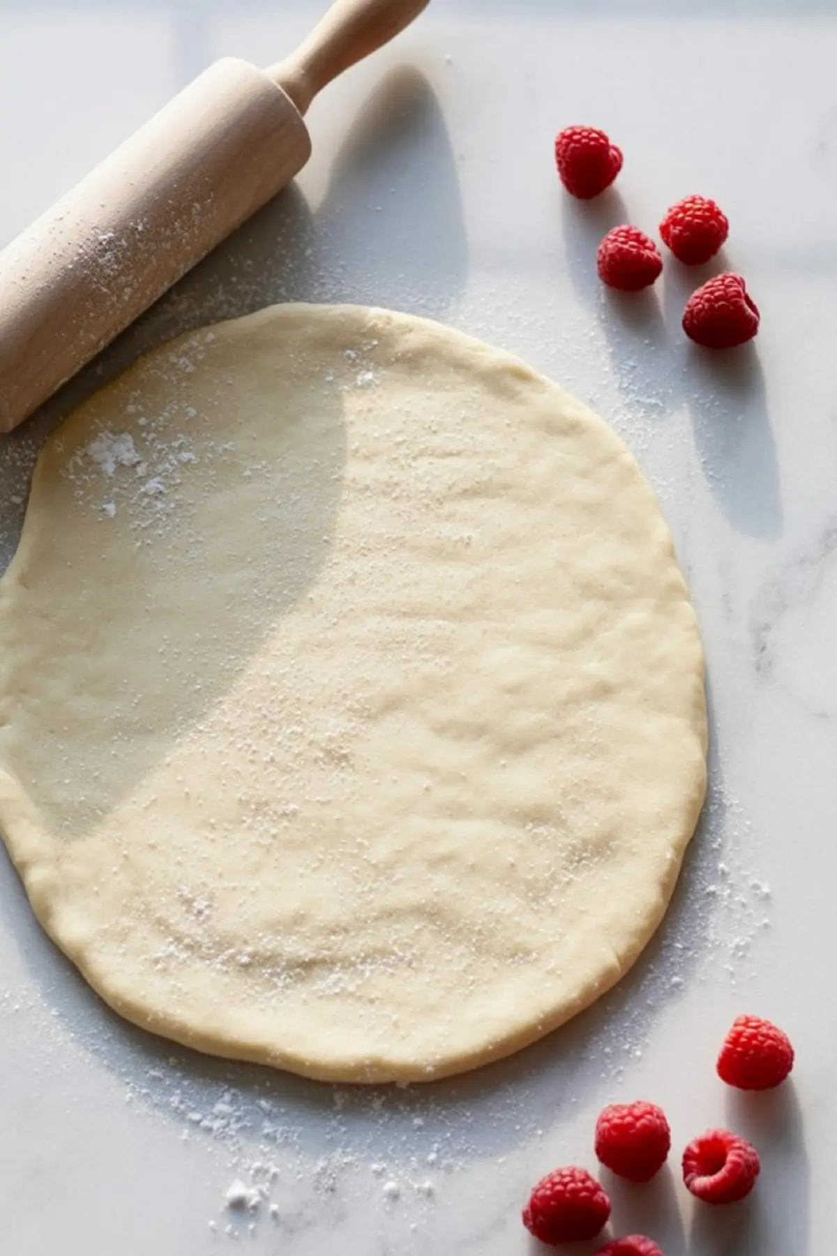 Rolled out sugar cookie dough dusted with flour, surrounded by scattered fresh raspberries and a wooden rolling pin on a light surface.