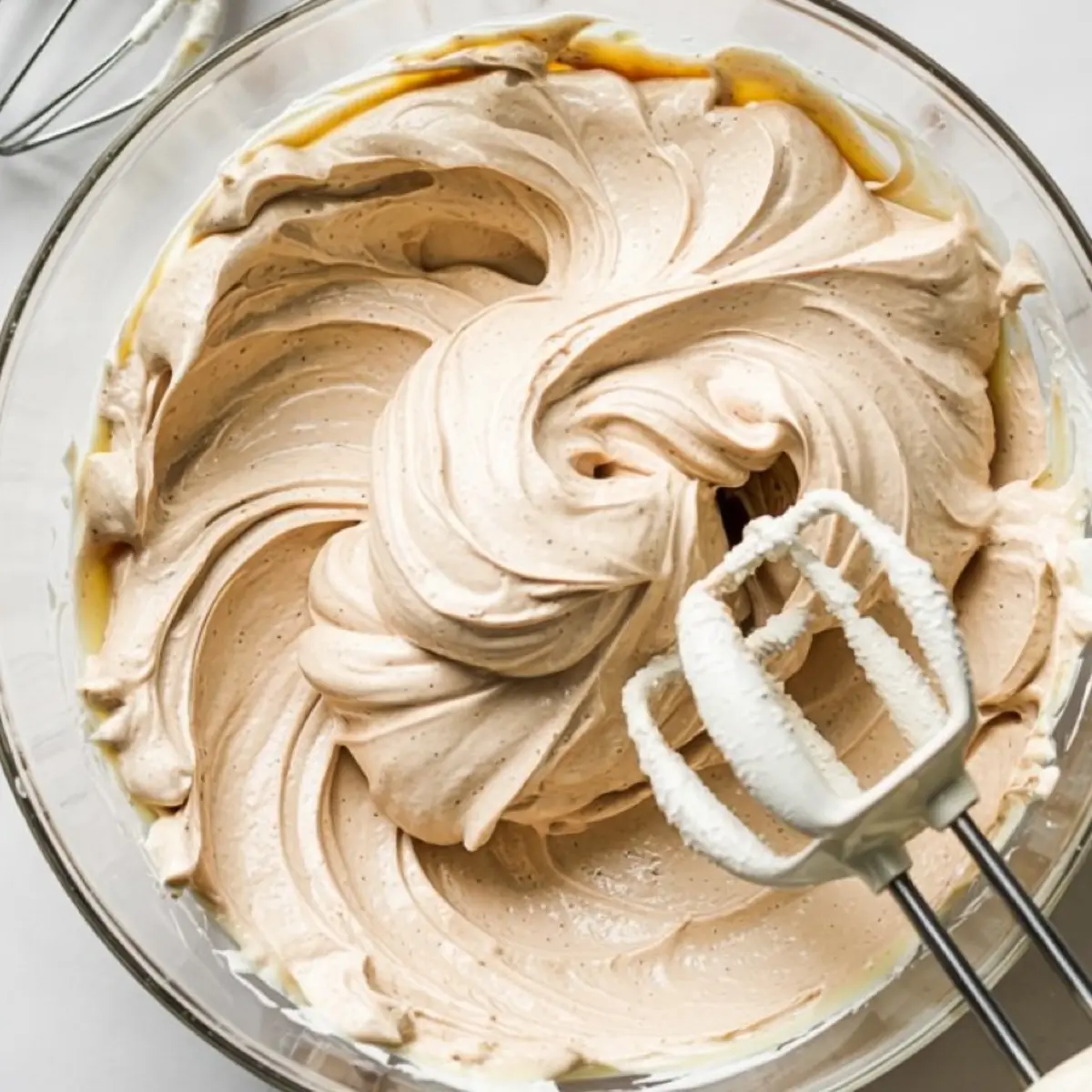 Creamy whipped frosting with a light caramel hue swirls smoothly in a glass bowl, with electric mixer beaters resting on the edge, showing a fully blended dairy-free frosting.