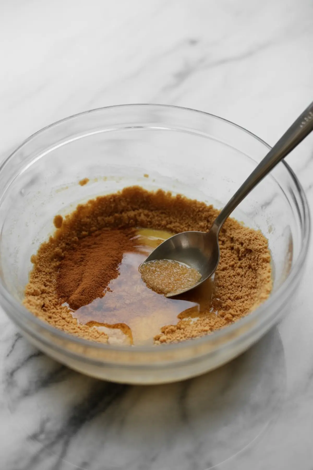 Cinnamon, brown sugar, and melted butter mixture in a glass bowl with a metal spoon, ready for graham cracker crust preparation.
