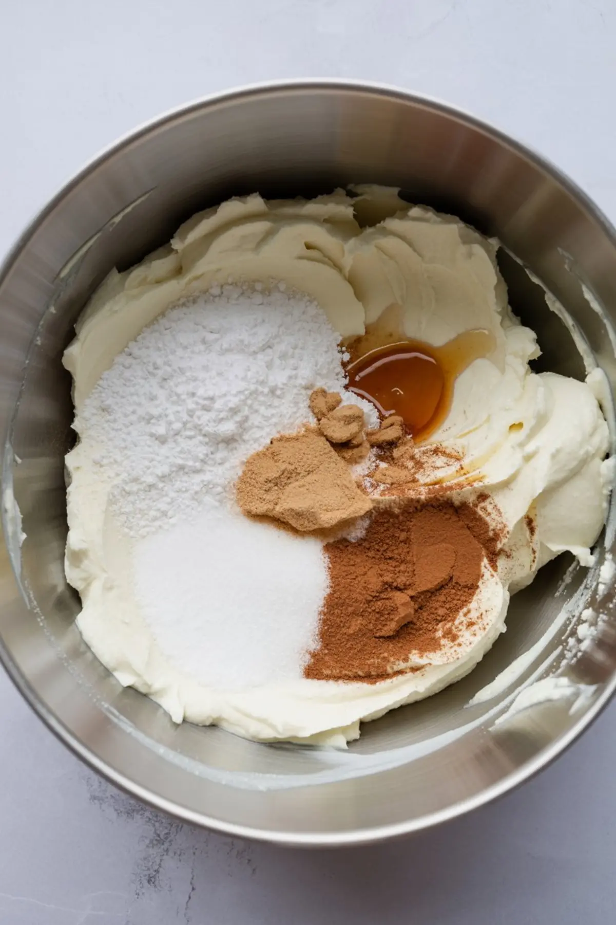 Stainless steel bowl filled with cream cheese, sugar, powdered sugar, cinnamon, ginger, and honey ready to be mixed for eggnog mousse.