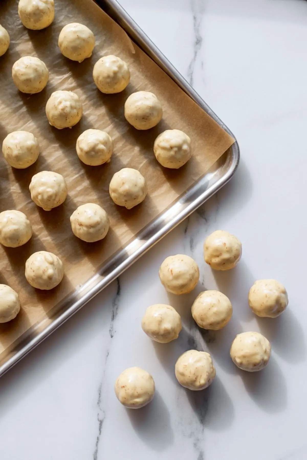 Round truffle balls on parchment paper-lined baking sheet and white marble surface, showcasing formed eggnog truffle centers before dipping.
