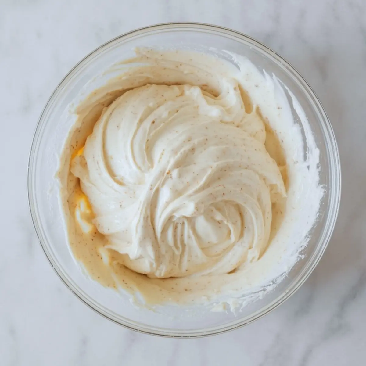 Smooth and fluffy eggnog cream cheese mixture in a glass bowl, swirled with visible spice flecks, set on a marble surface.
