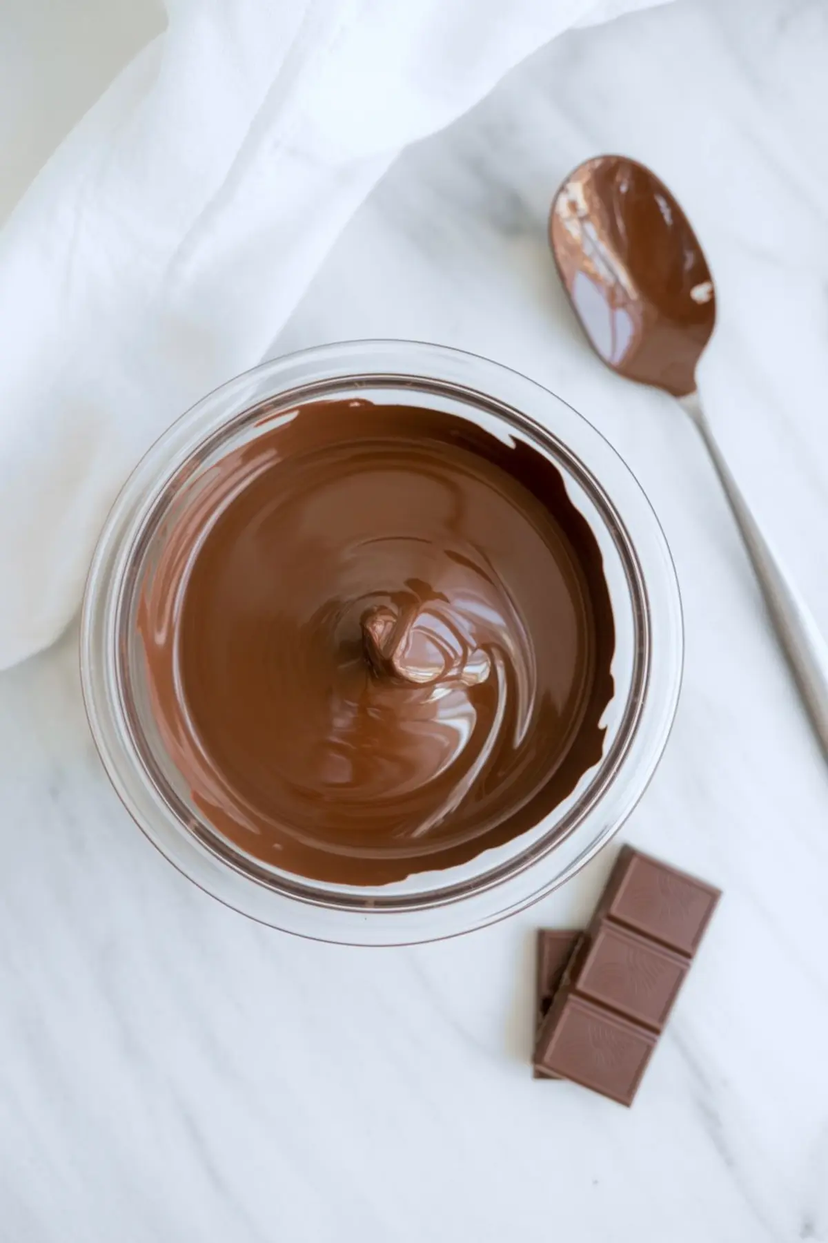 Glossy bowl of melted milk chocolate with spoon and chocolate squares nearby, ready for truffle coating on a white marble background.
