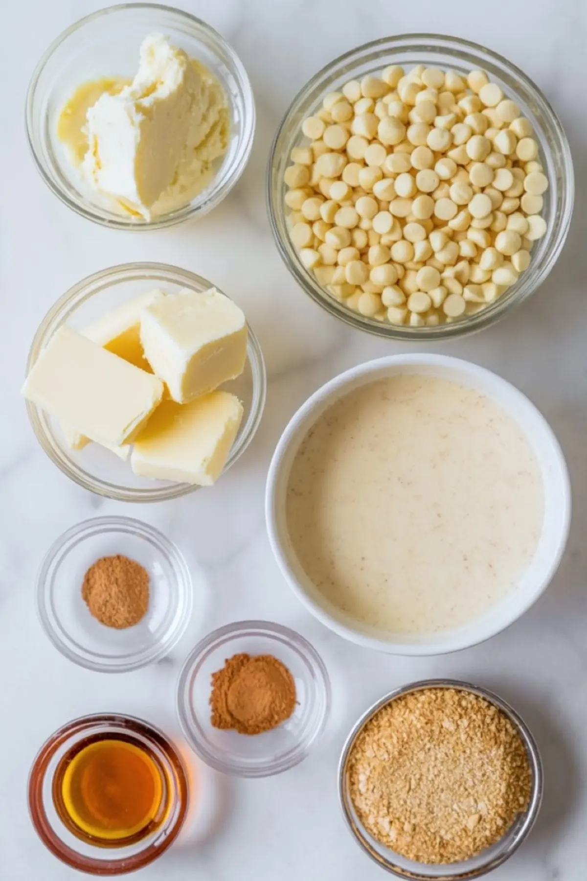 Flat lay of eggnog truffle ingredients including white chocolate chips, butter, cream cheese, graham cracker crumbs, cinnamon, nutmeg, eggnog, and vanilla extract in small glass bowls.
