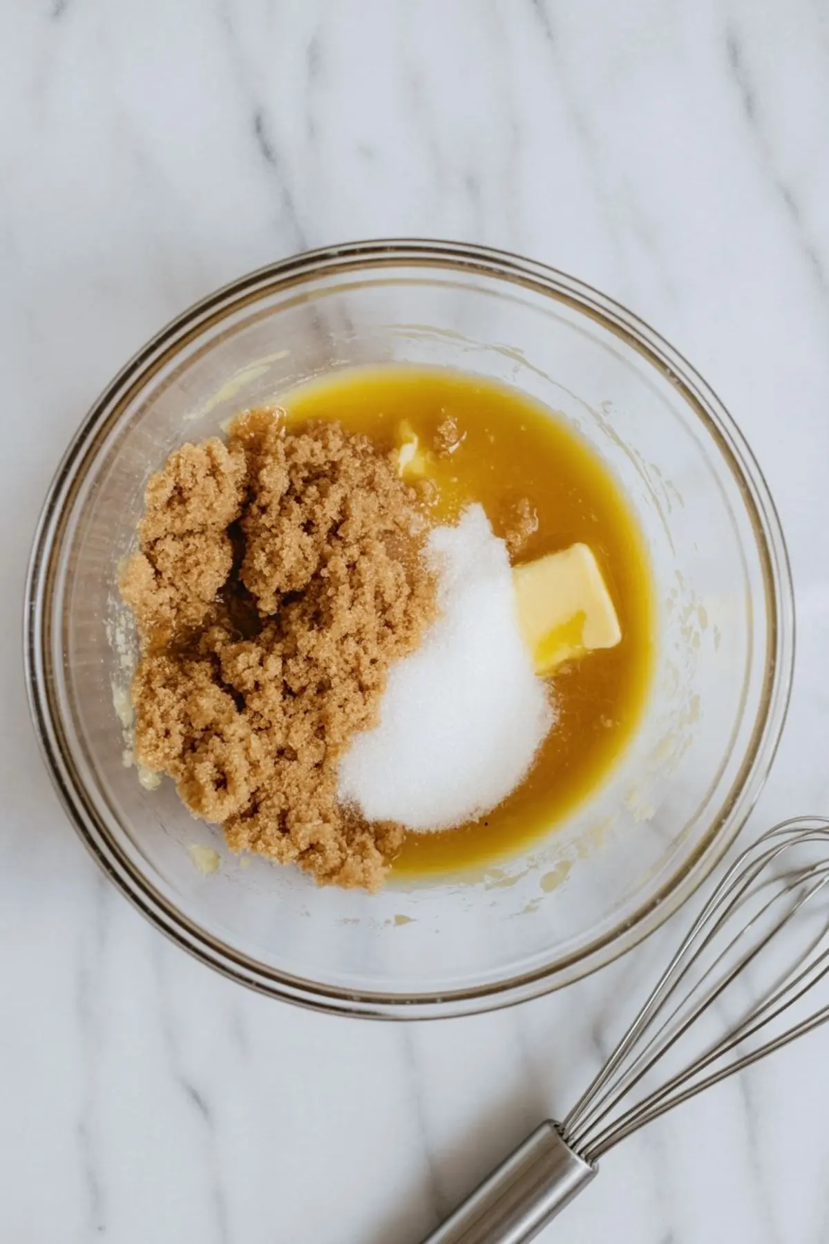 A mixing bowl with melted butter, brown sugar, and white sugar partially blended together, with a metal whisk resting beside it on a marble countertop.