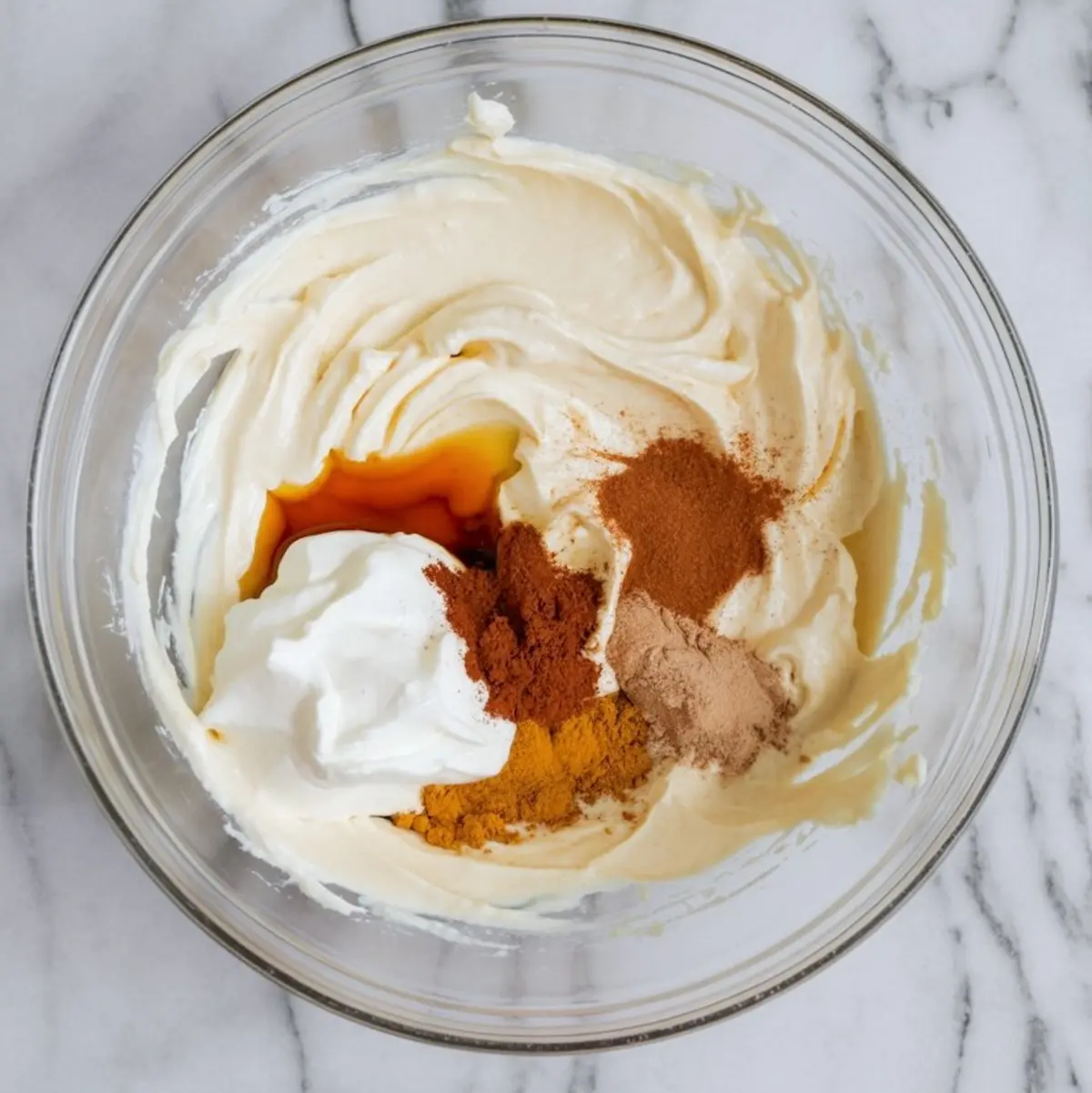 A mixing bowl displays a blend of cream cheese mixture with swirls of vanilla extract, whipped cream, and spices including cinnamon, ginger, and nutmeg on top, capturing the start of a spiced holiday dessert base.
