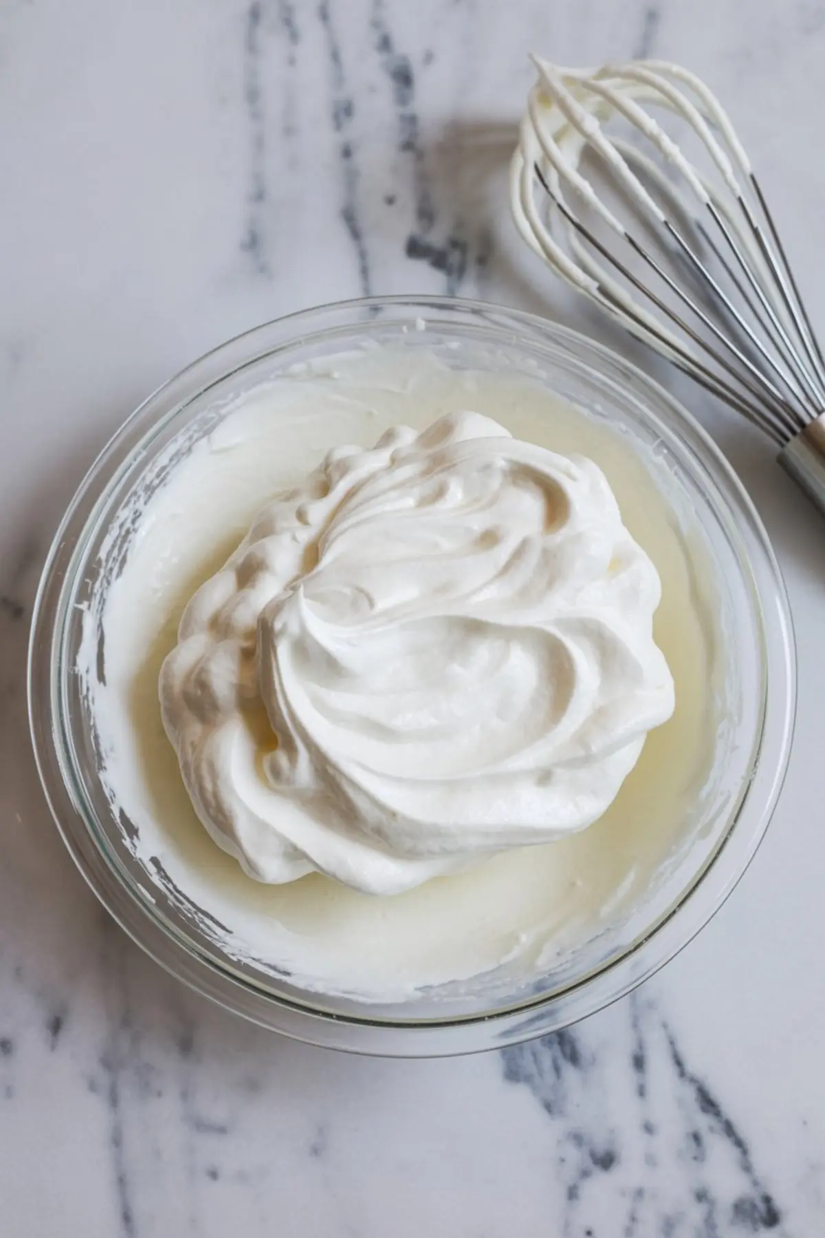A glass bowl filled with stiff peaks of freshly whipped cream sits on a marble surface next to a metal whisk coated with cream, showcasing smooth and airy texture ideal for dessert toppings or fillings.
