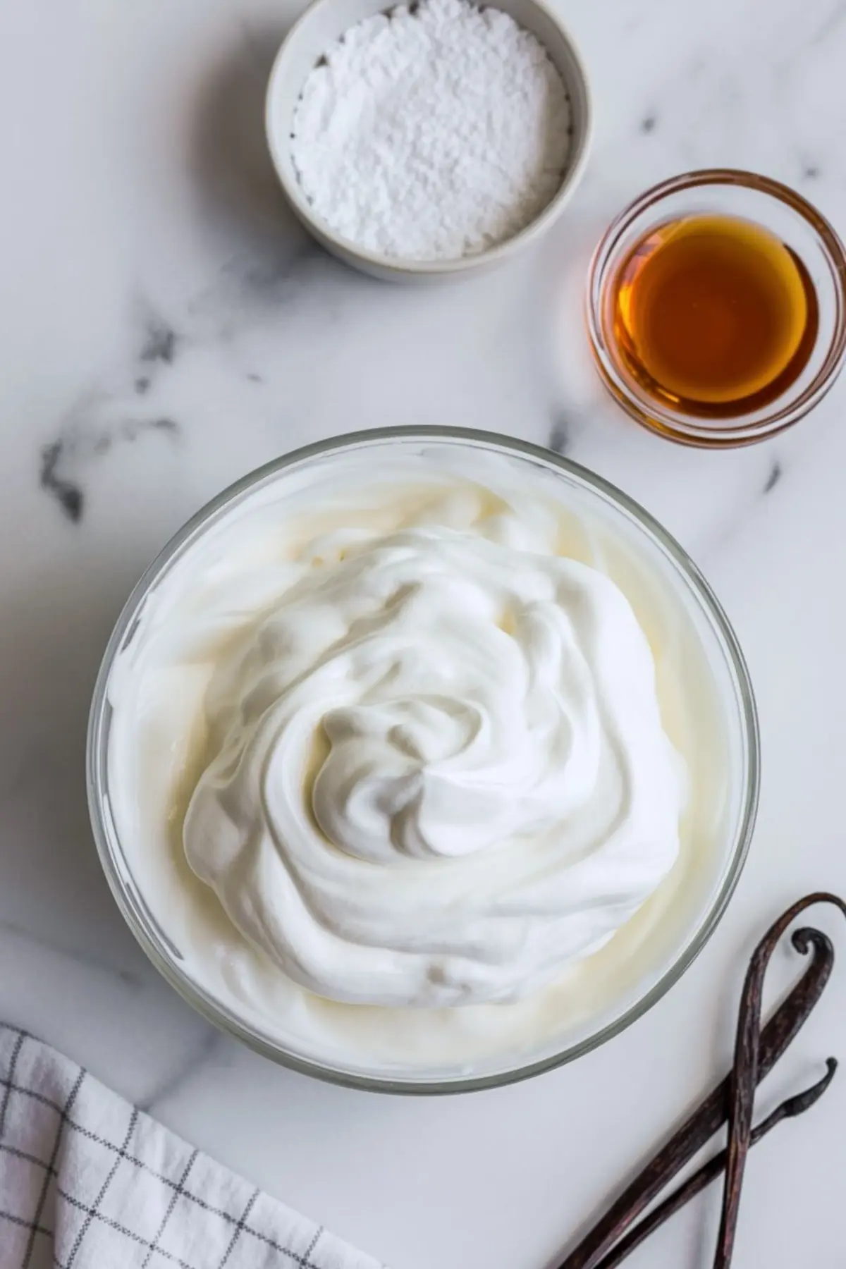 A bowl of fluffy whipped cream is positioned beside a small bowl of powdered sugar and a glass bowl of vanilla extract, with vanilla beans and a striped cloth nearby on a marble counter, highlighting key ingredients for homemade frosting or mousse.

