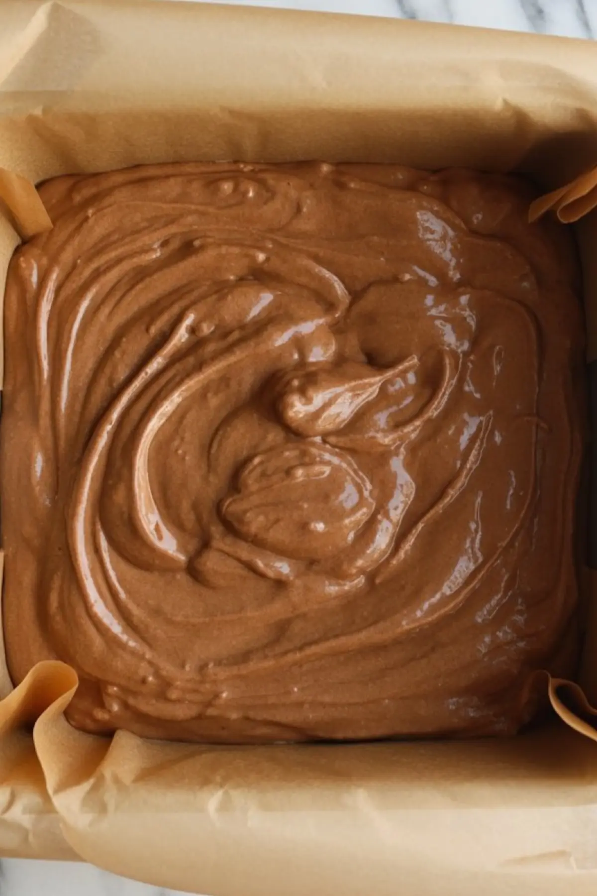 Square baking dish lined with parchment paper filled with freshly poured gingerbread fudge batter, showcasing a thick, swirled texture ready for setting.