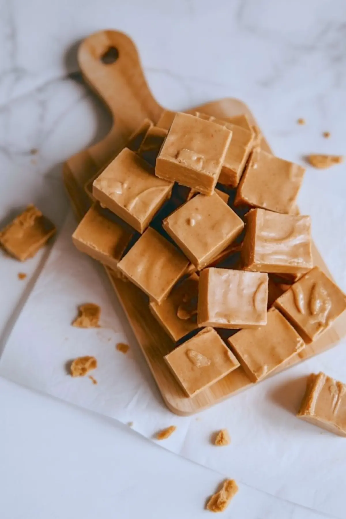 Stack of creamy gingerbread fudge squares arranged on a wooden cutting board, with smooth texture and golden-brown color, surrounded by small broken fudge crumbs on a marble surface.