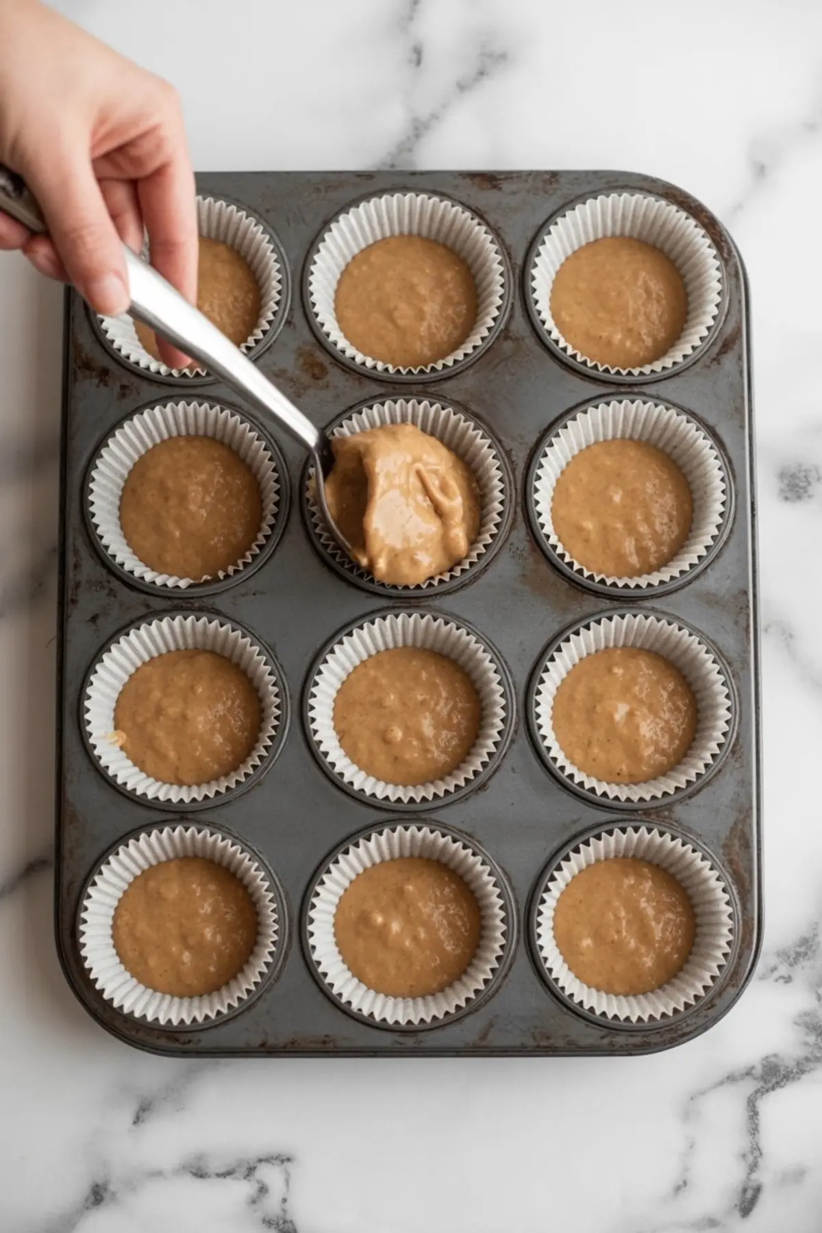 A muffin pan lined with white cupcake liners, each filled with gingerbread batter as a hand uses a cookie scoop to portion the mixture.
