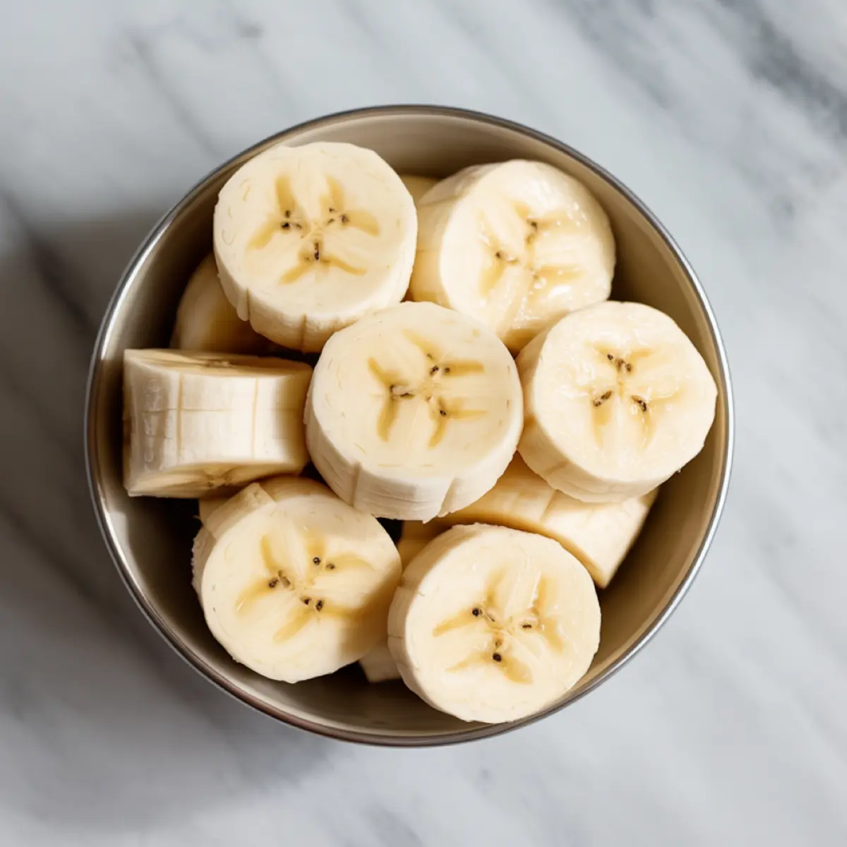 Metal bowl holds neatly sliced frozen banana rounds on cool white marble, ready to thicken a creamy vegan smoothie.