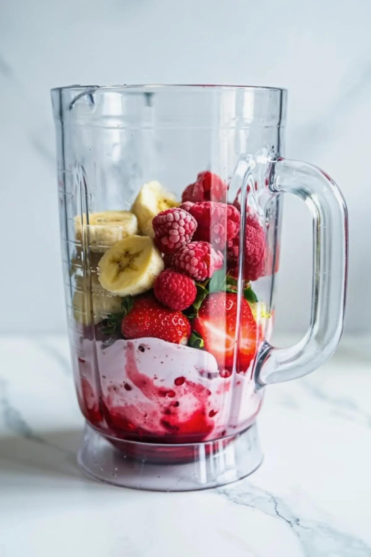 Fresh banana slices, strawberries, and raspberries layered over pink yogurt in a clear blender pitcher, ready to blend into a berry smoothie.
