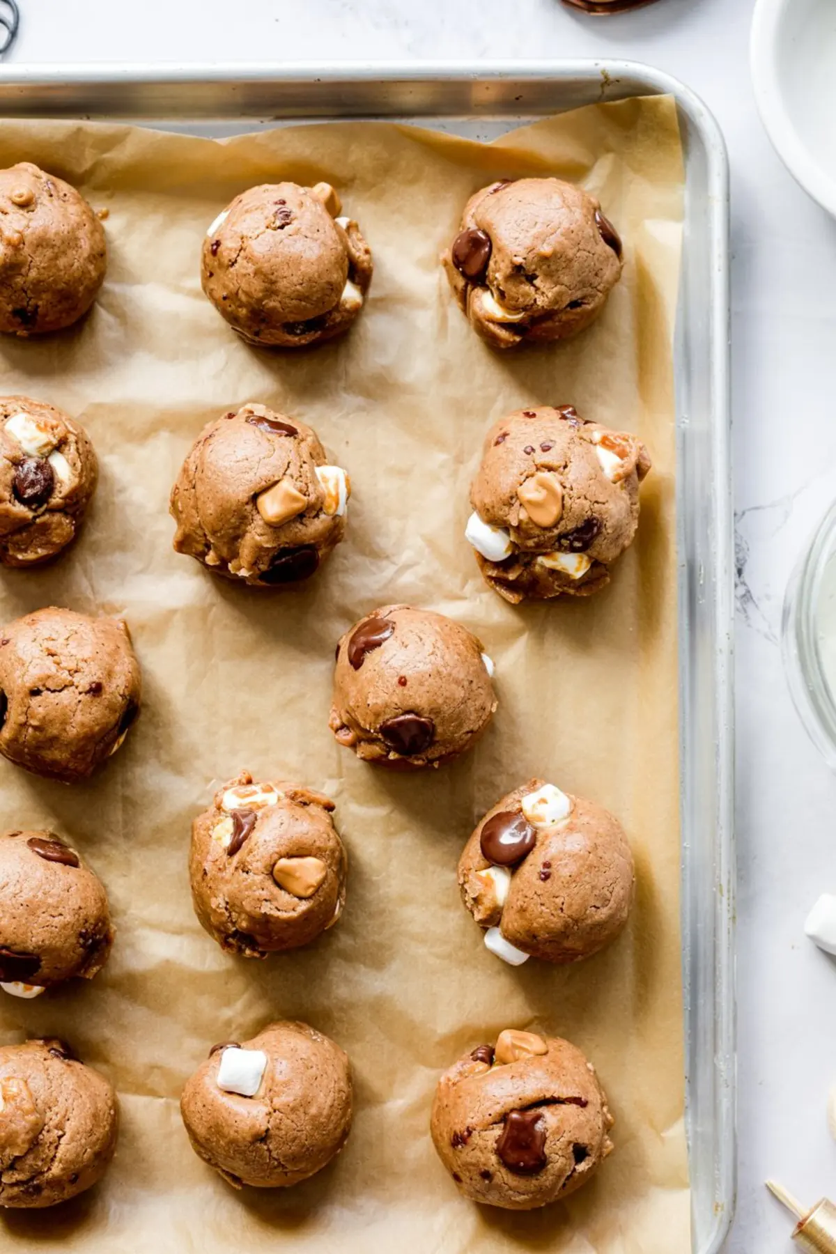 Raw cookie dough balls packed with chocolate chunks, caramel bits, and mini marshmallows arranged on a parchment-lined baking sheet.