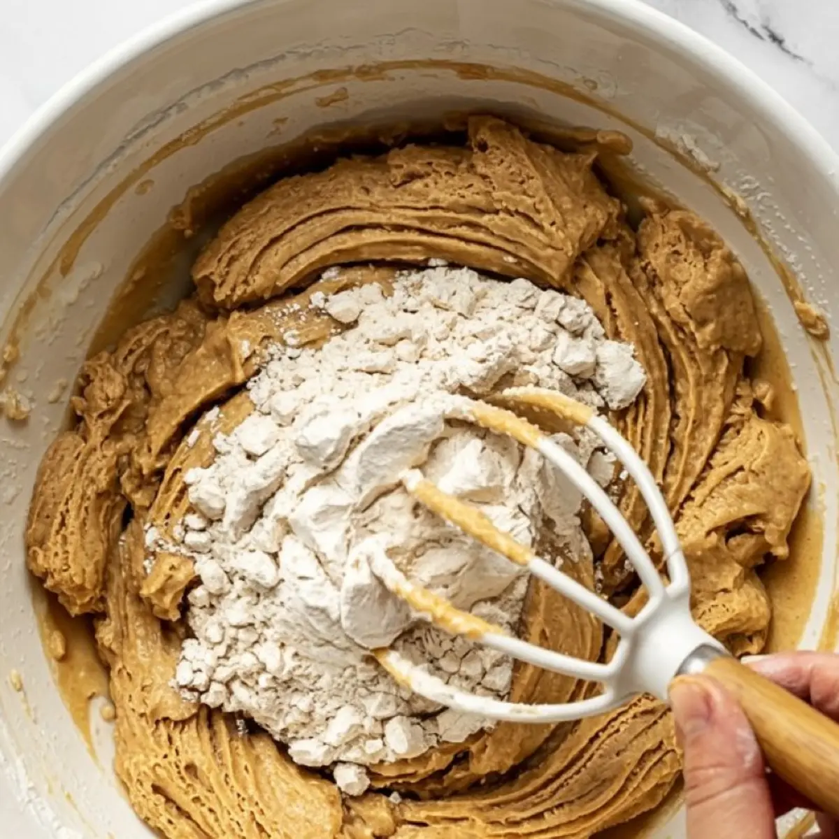 Partially mixed cookie dough with a layer of flour on top being blended using a hand mixer in a white bowl.
