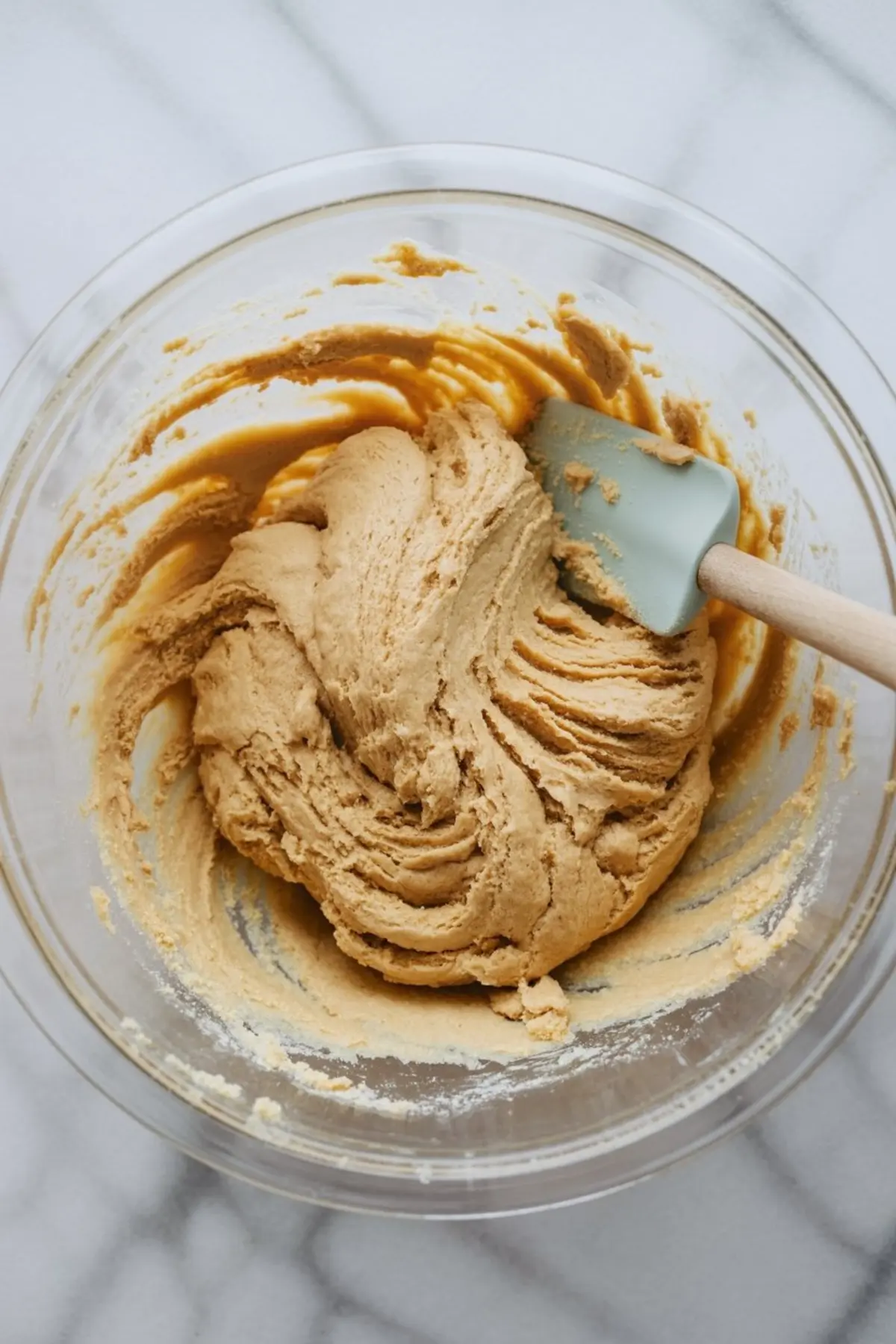 Thick, creamy cookie dough with a golden-brown hue mixed in a glass bowl, being stirred with a light blue silicone spatula, capturing the maple brown sugar cookie batter texture.
