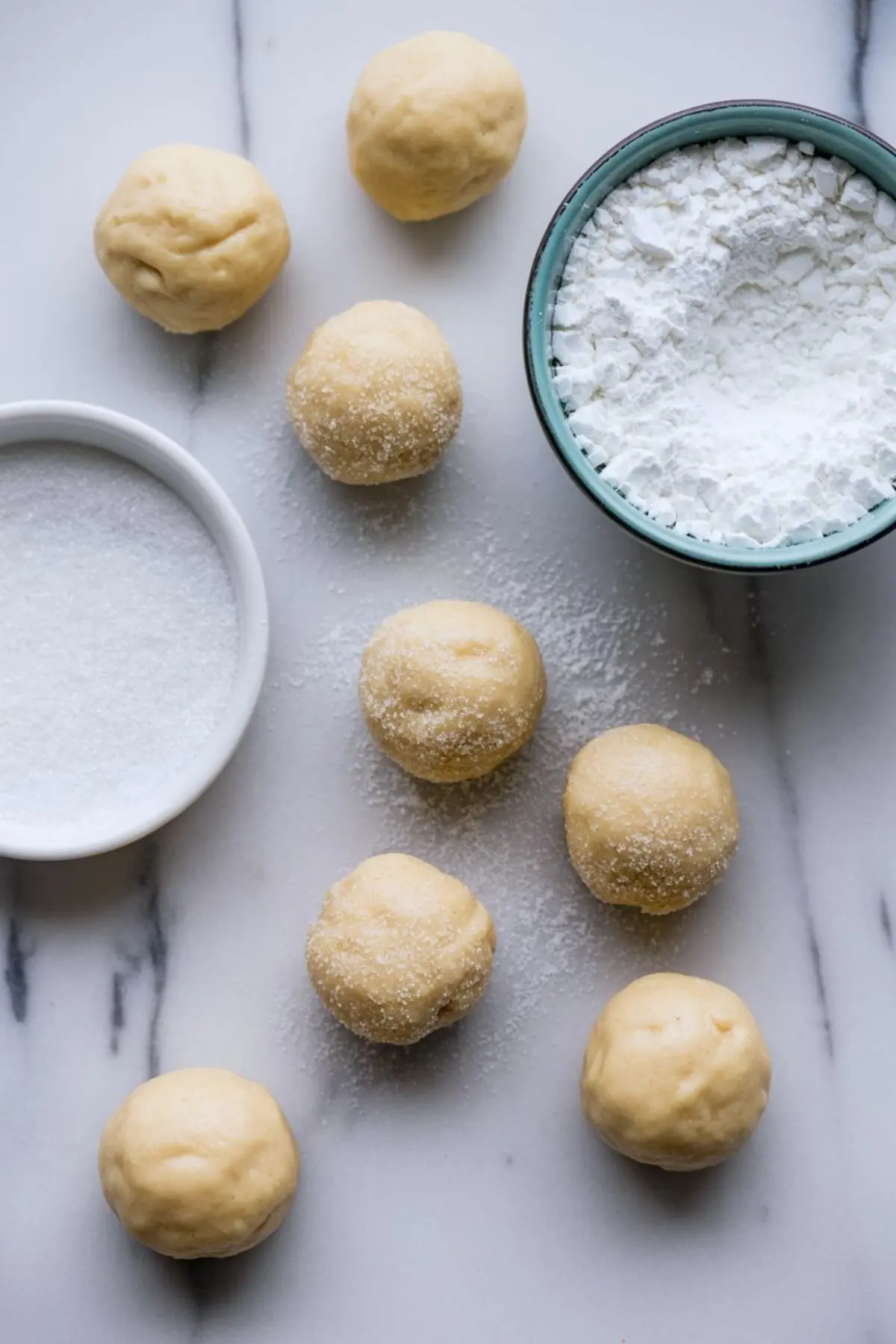 Rolled cookie dough balls on a marble surface, some coated in granulated sugar, placed next to a bowl of powdered sugar, ready for baking maple crinkle cookies.
