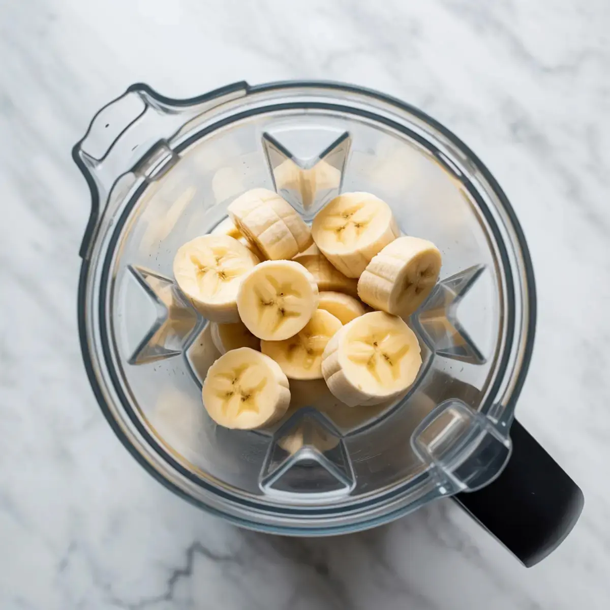 Fresh banana slices arranged inside a high-speed blender pitcher, placed on a white marble surface, ready for smoothie preparation.
