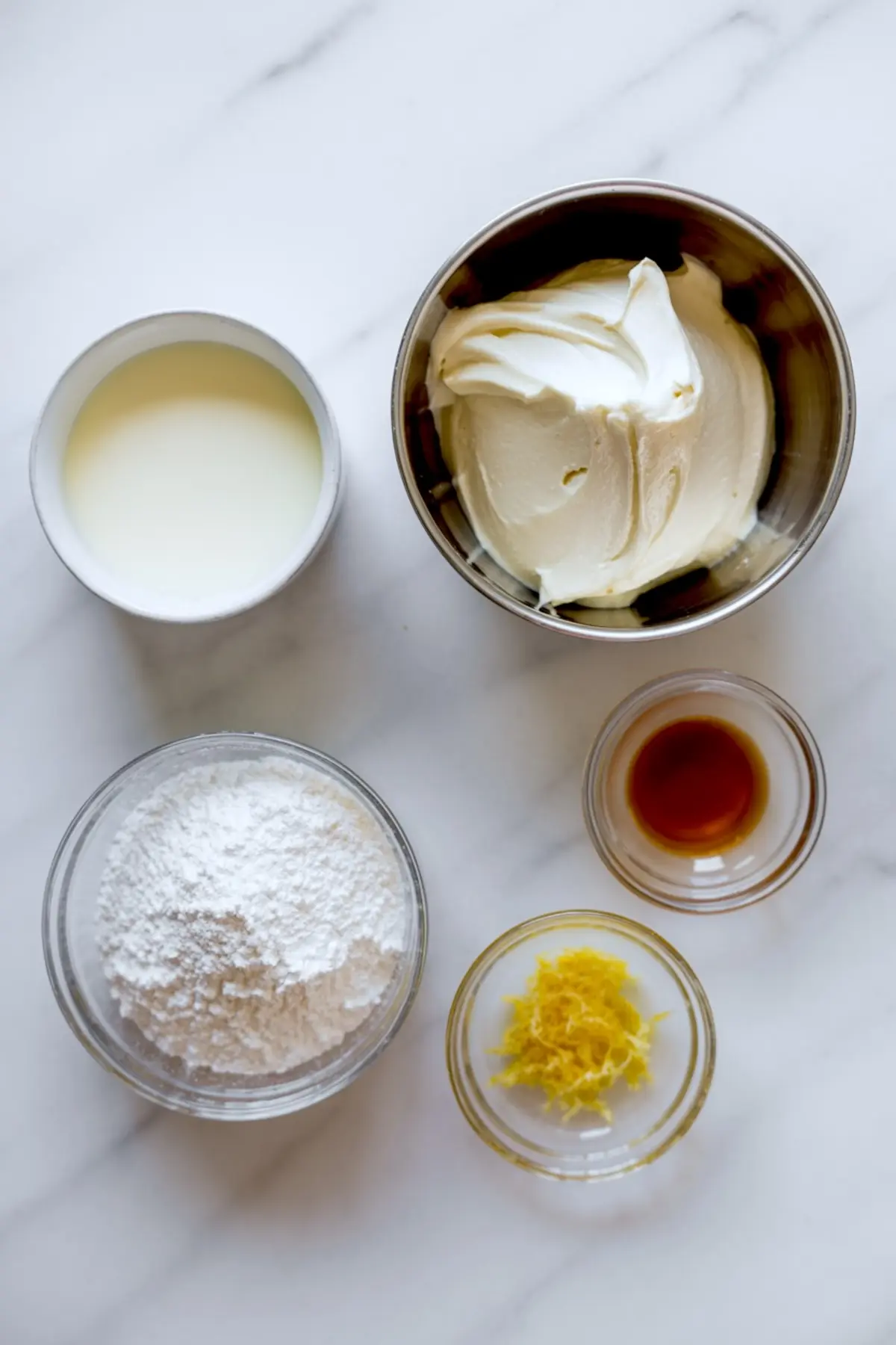 Overhead view of mascarpone frosting ingredients including powdered sugar, vanilla extract, lemon zest, mascarpone cheese, and heavy cream in individual bowls on a white marble countertop.