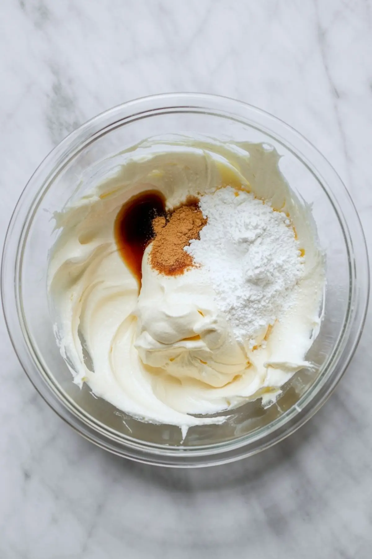 Whipped mascarpone cheese in a glass bowl with added vanilla extract, ground cinnamon, and powdered sugar, ready to be mixed into frosting.