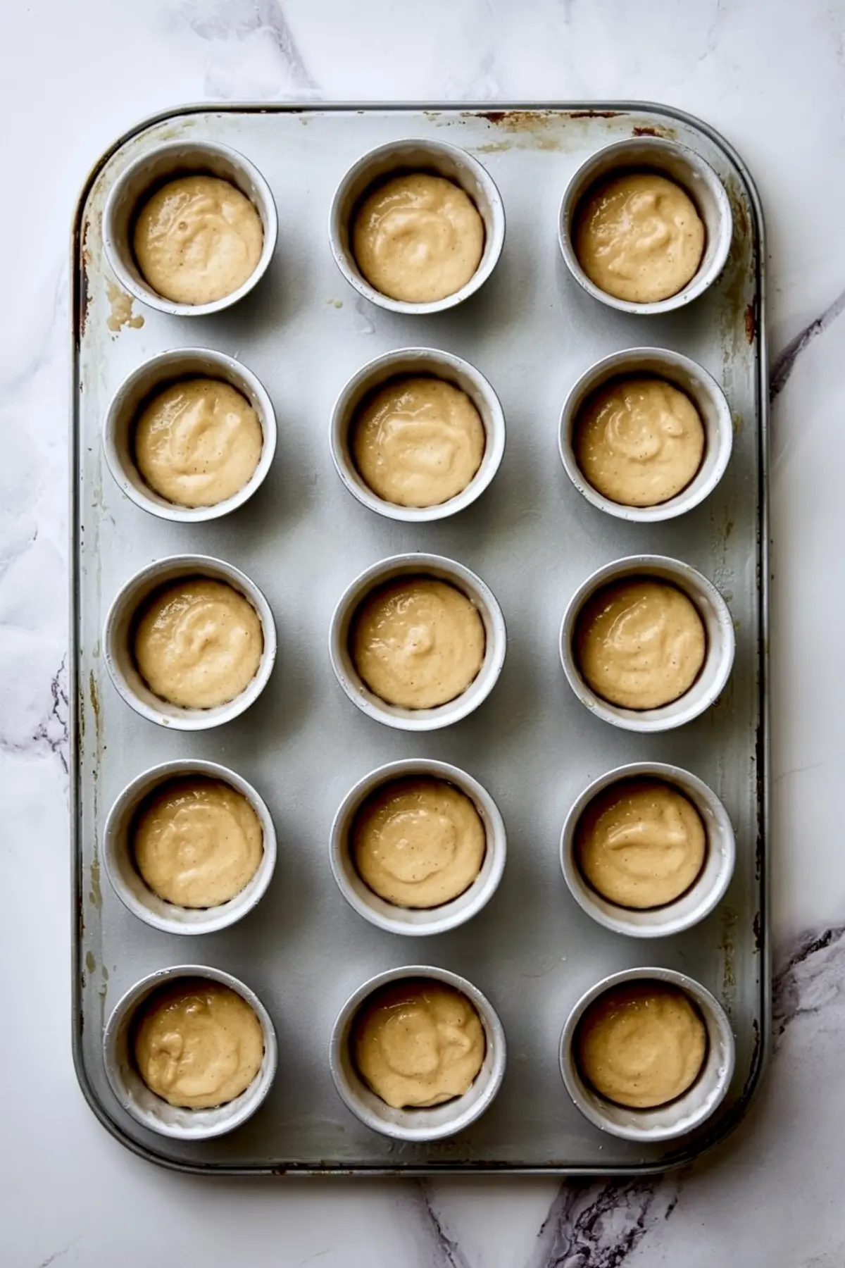 Muffin tin filled with uncooked cupcake batter, evenly portioned in each cup on a lightly worn metal tray.