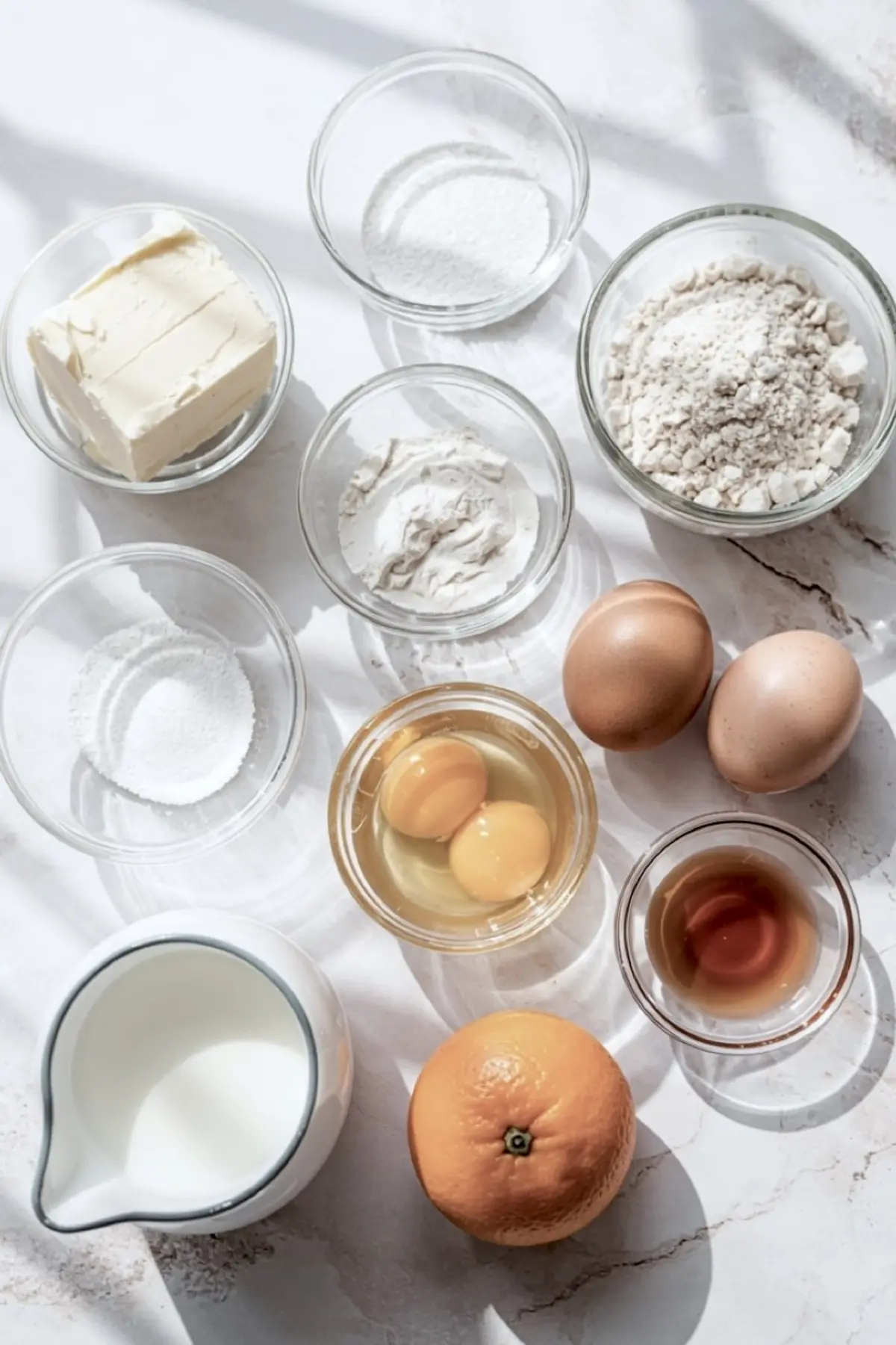 Flat lay of baking ingredients in glass bowls, including flour, sugar, butter, eggs, vanilla extract, baking powder, milk, and a fresh orange on a sunlit marble surface.