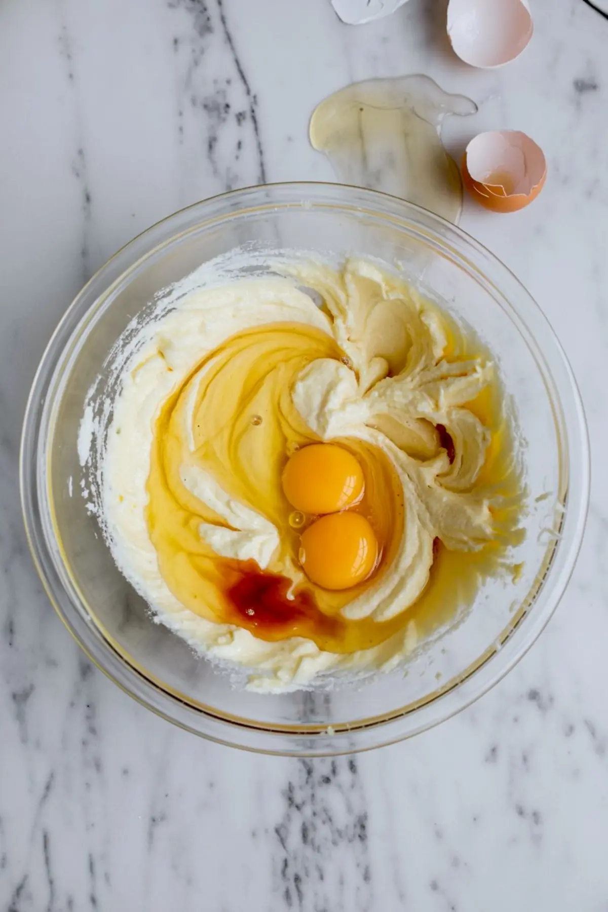 Creamed butter and sugar mixture in a glass bowl with eggs and vanilla extract added on top, prepared on a marble surface for chocolate cake baking.
