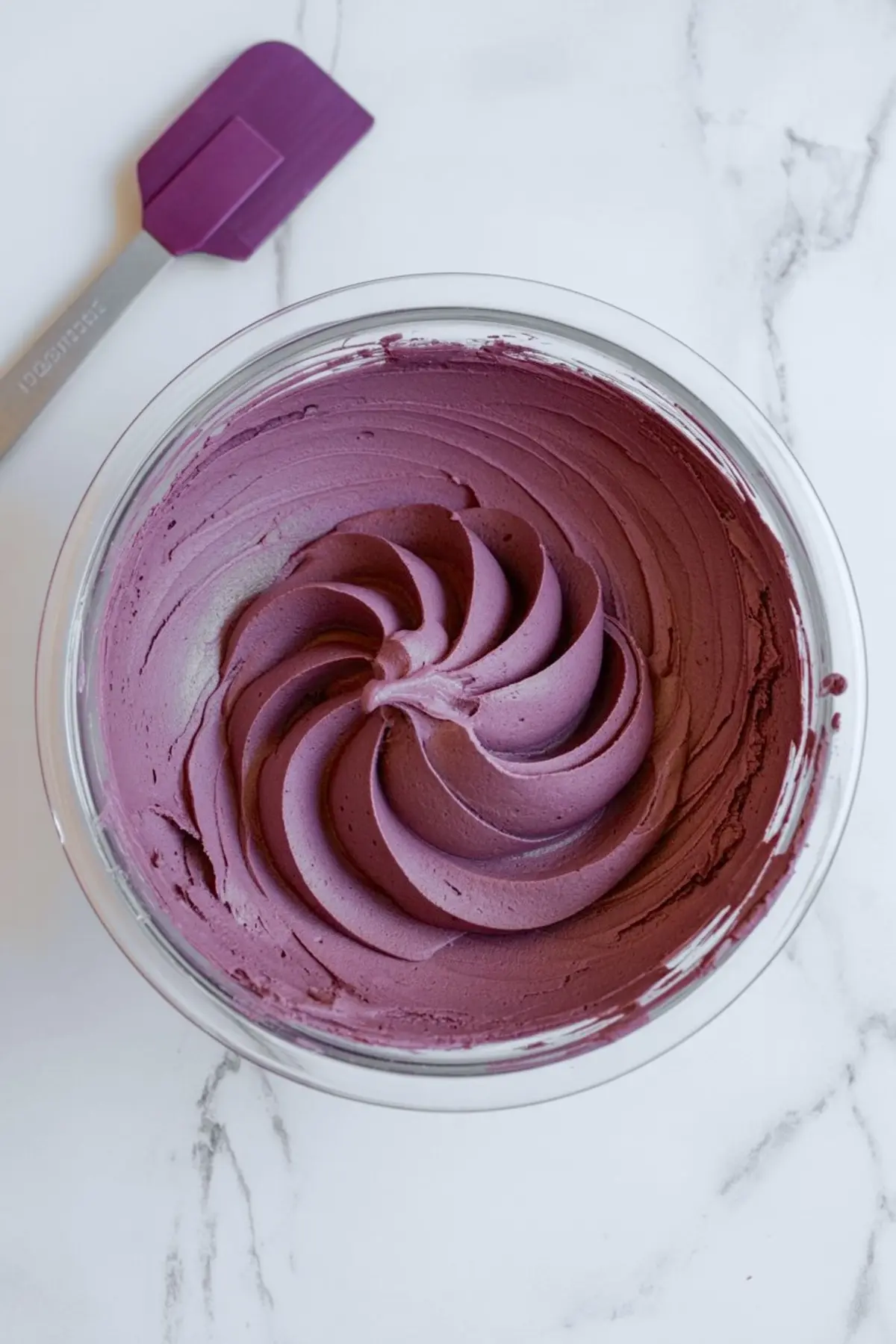 Smooth and creamy purple buttercream frosting swirled in a glass bowl with a matching purple spatula beside it, ready for cake decorating.
