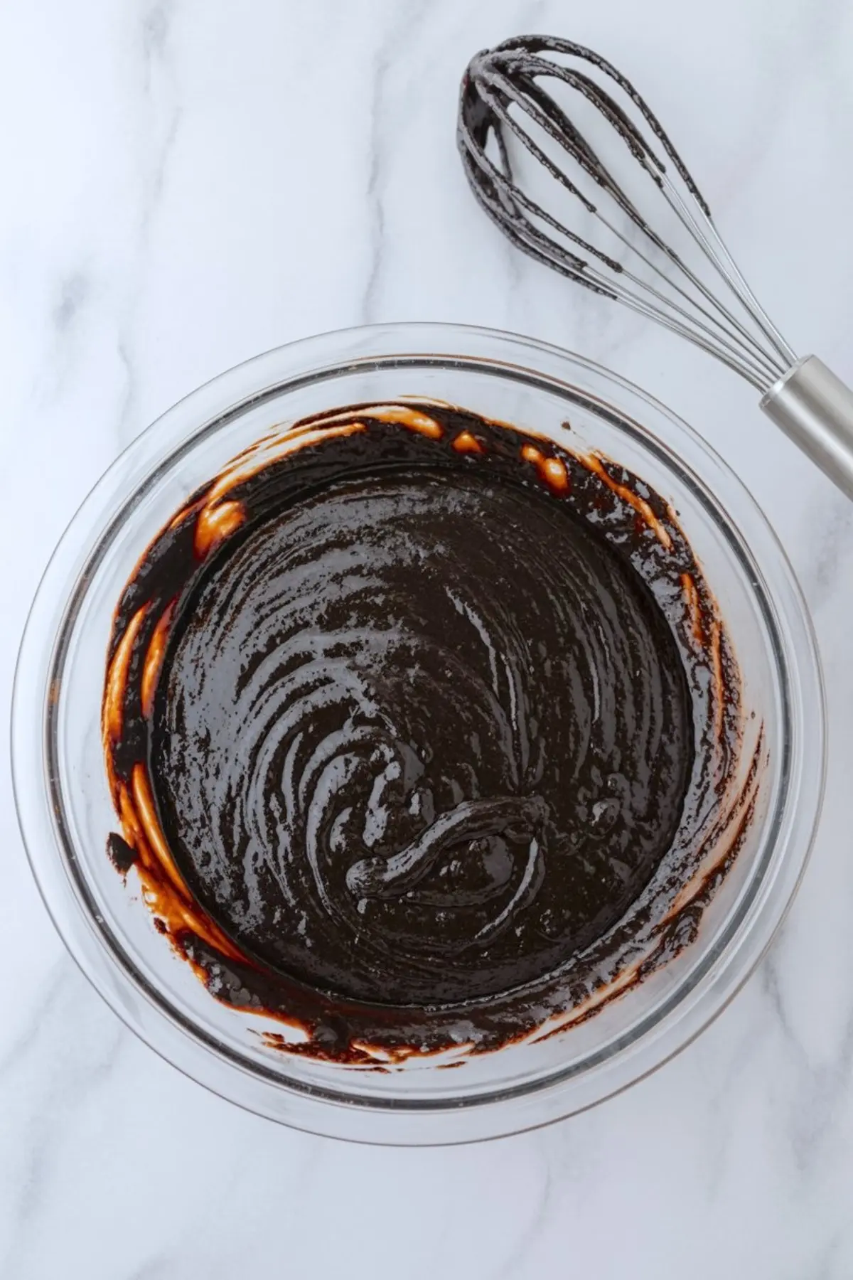 Glossy and thick chocolate cake batter in a glass mixing bowl with a whisk coated in batter placed next to it on a marble surface.
