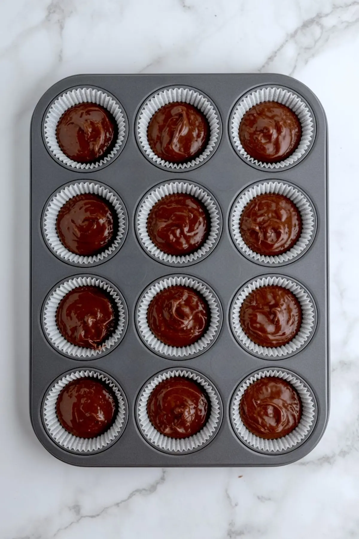 Chocolate cupcake batter scooped into a 12-cup muffin tin lined with white paper liners, ready to bake on a marble counter.