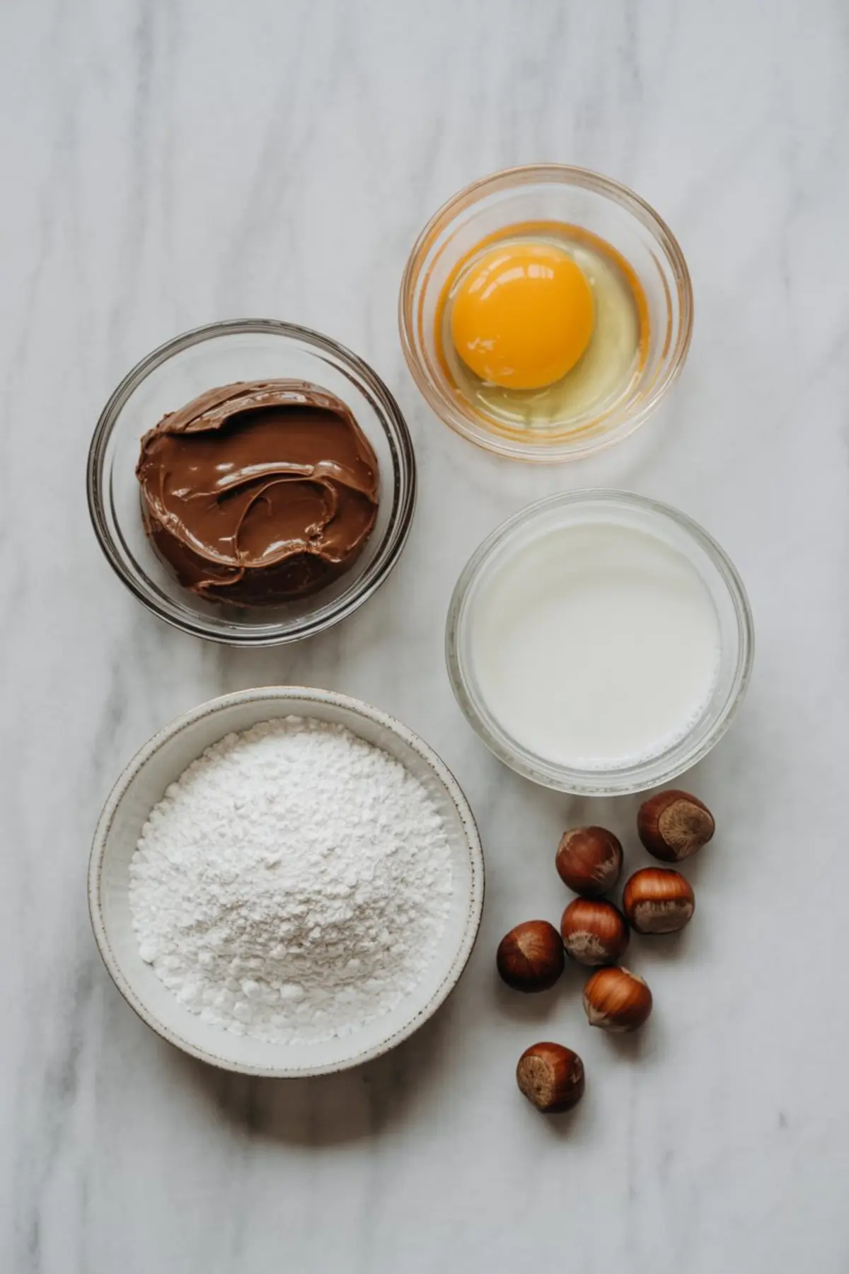 Flat lay of Nutella Christmas tree ingredients including a raw egg, milk, powdered sugar, chocolate hazelnut spread, and whole hazelnuts in clear and ceramic bowls on a light marble surface.
