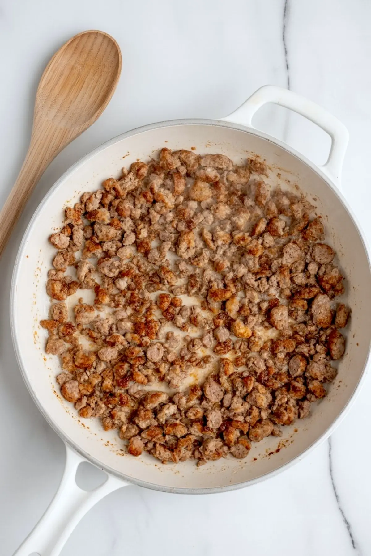 Crispy browned sausage crumbles cooking in a white skillet with a wooden spoon on the side, prepared as the first step for a homemade breakfast casserole recipe.
