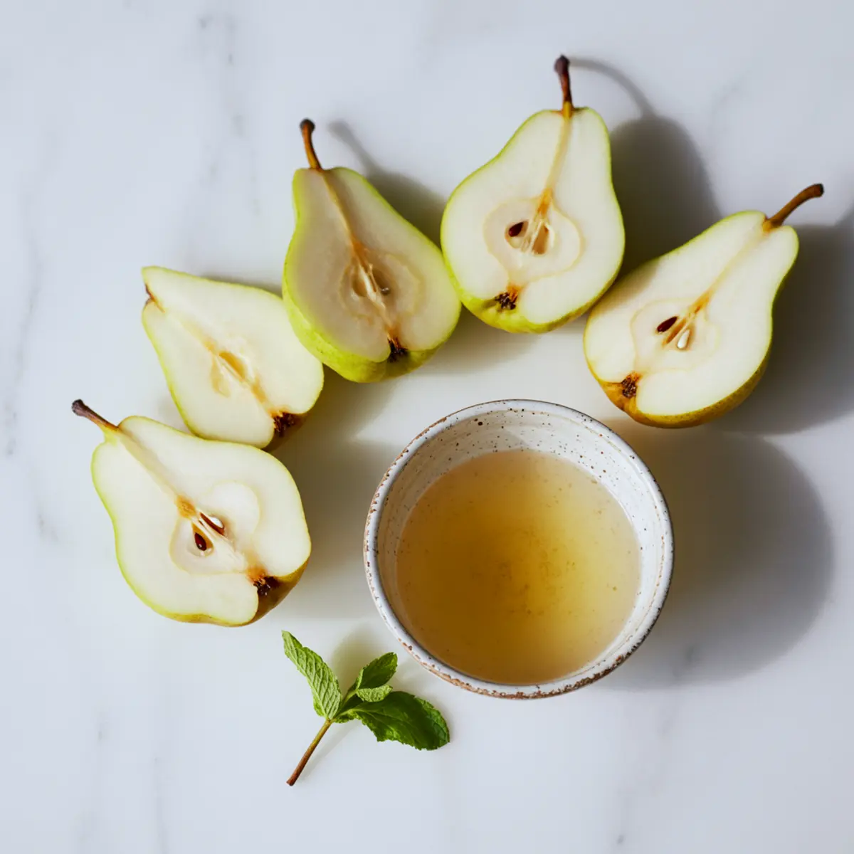 Halved green pears arranged in a semi-circle around a ceramic bowl of golden pear juice with a fresh mint sprig on a white marble surface.