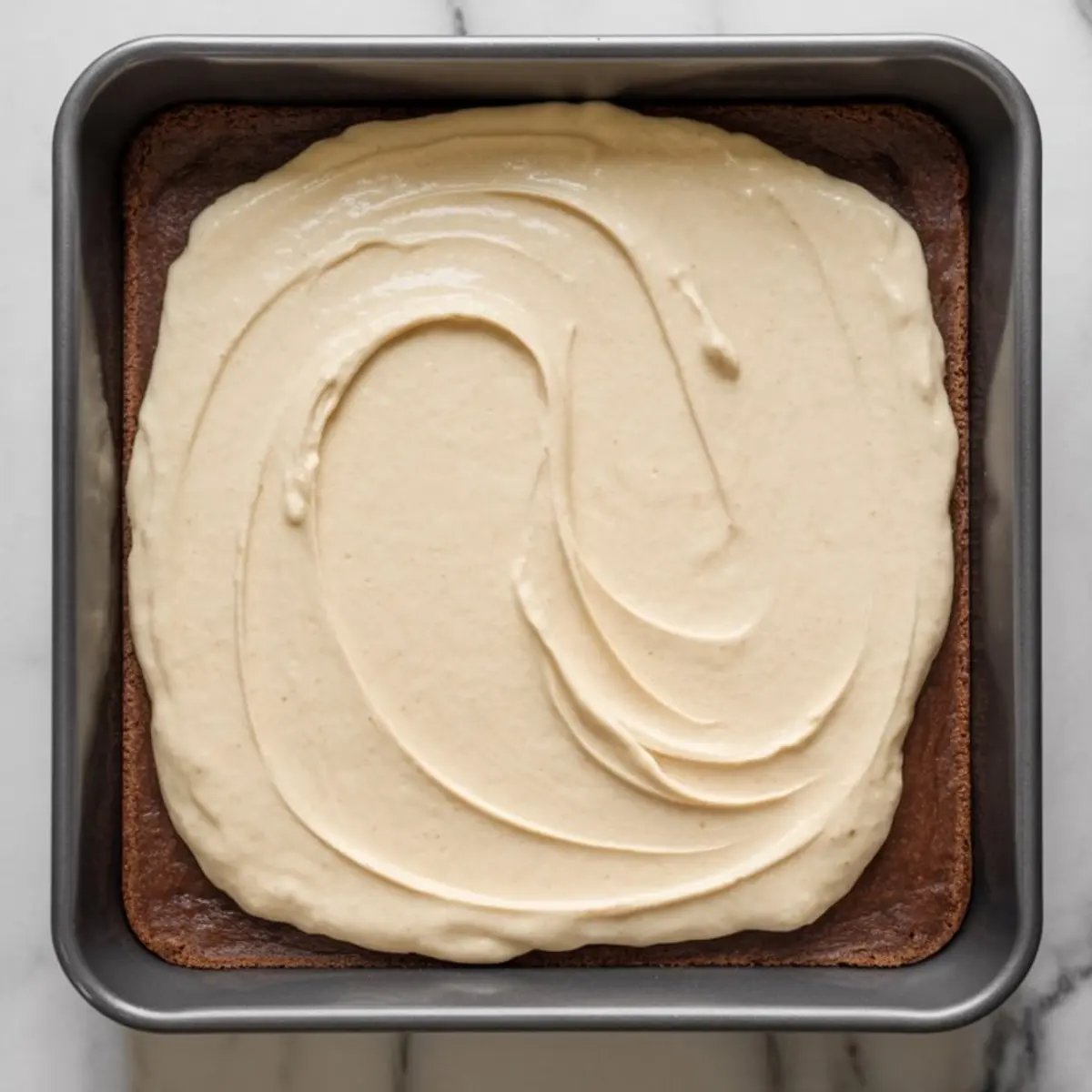 Smooth cheesecake batter spread evenly over a baked chocolate crust inside a square metal pan, ready for baking peppermint bark cheesecake bars.
