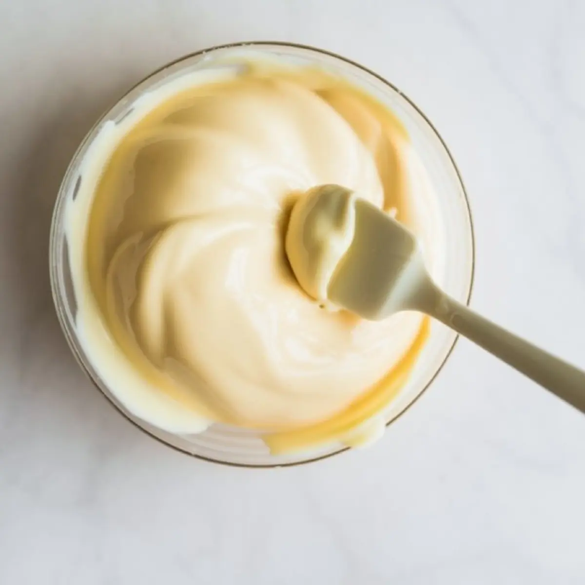 Thick and creamy cheesecake batter being mixed with a rubber spatula in a clear glass bowl, showing smooth texture and consistency.
