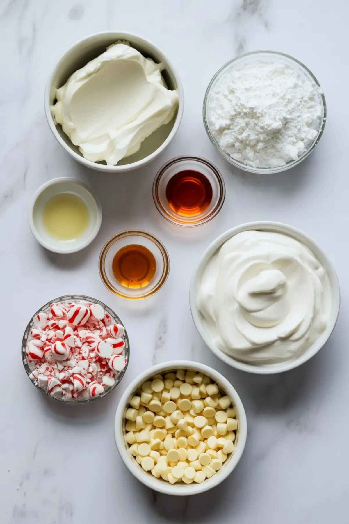 Flat lay of peppermint cheesecake dip ingredients including whipped topping, cream cheese, powdered sugar, peppermint candies, white chocolate chips, vanilla extract, and other flavorings in small bowls on a marble background.
