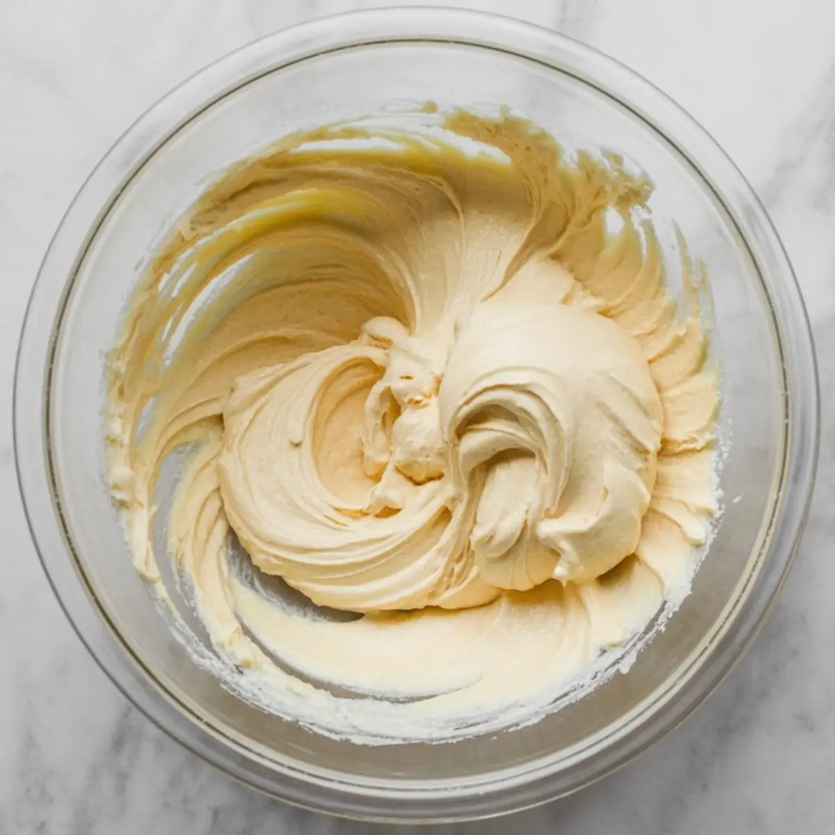 Glass bowl of smooth, pale yellow cheesecake mixture swirled into a creamy texture, ready for the next step in the dessert recipe.

