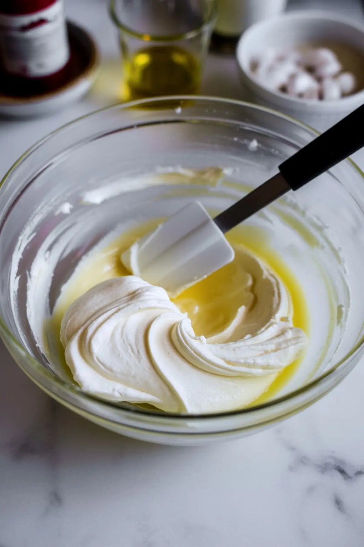 Partially mixed peppermint cheesecake base in a glass bowl with whipped topping being folded in using a spatula, surrounded by blurred ingredients in the background.
