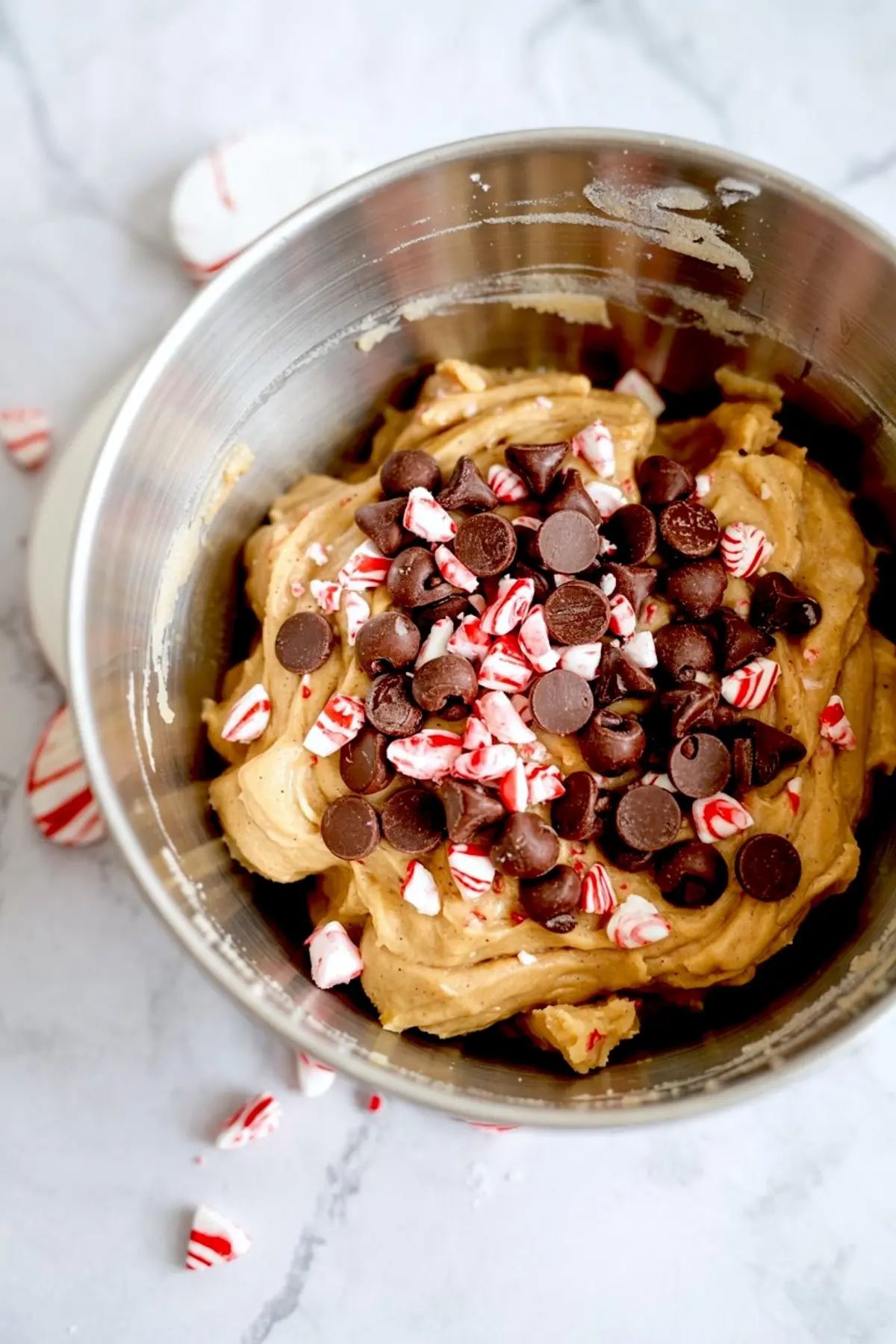 Cookie dough in a metal mixing bowl topped with crushed peppermint candy and chocolate chips, ready for baking festive holiday treats.

