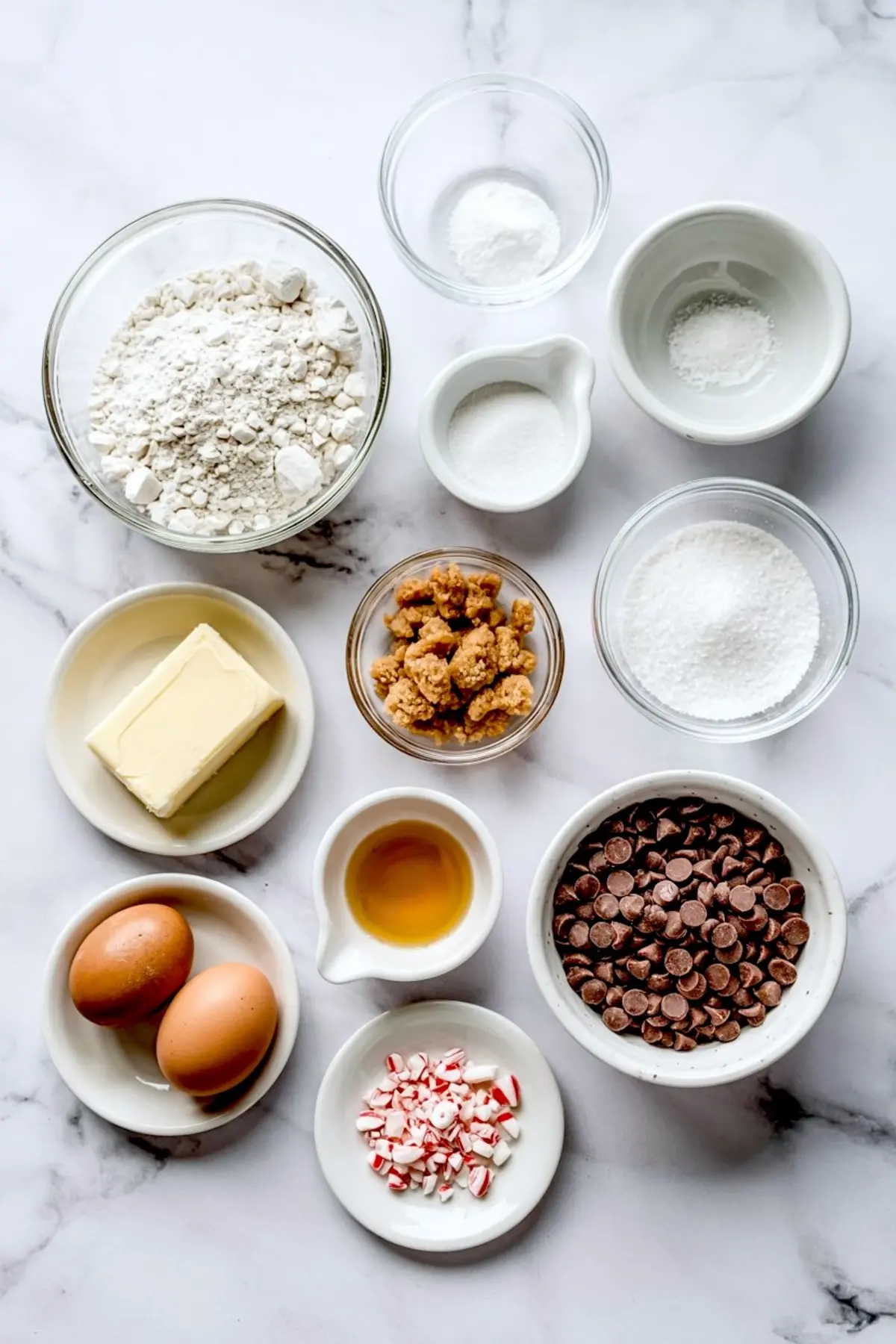 Flat lay of baking ingredients including flour, butter, sugar, eggs, vanilla extract, chocolate chips, and crushed peppermint in small bowls on a marble surface.
