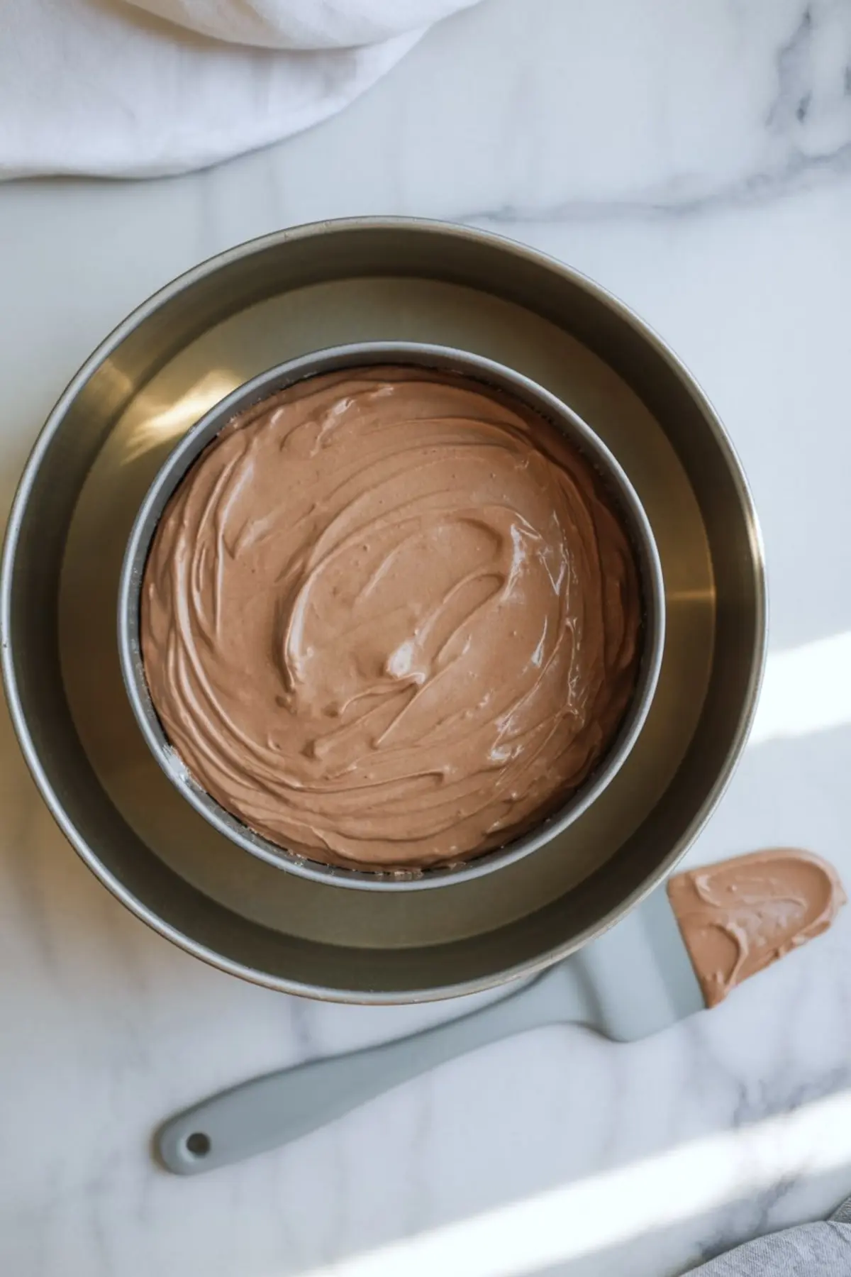 A baking pan filled with smooth mocha cheesecake batter, resting inside a water bath setup for even baking. The silky texture of the chocolate mixture is evenly spread in the pan, placed on a light marble countertop with a spatula nearby.
