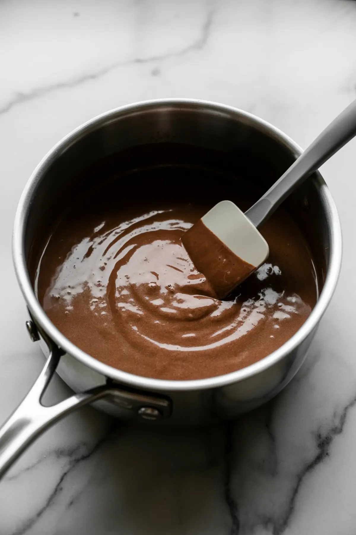 Glossy chocolate pudding mixture being stirred with a silicone spatula inside a stainless steel saucepan on a marble countertop.
