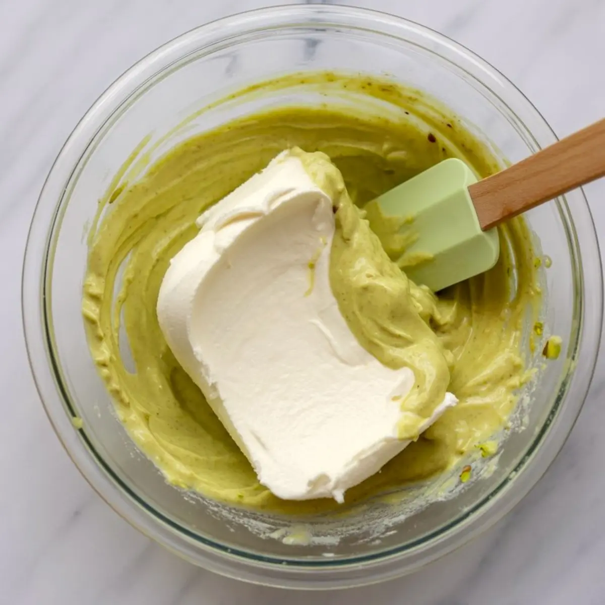 Glass bowl showing thick pistachio filling being blended with whipped cream using a green silicone spatula.
