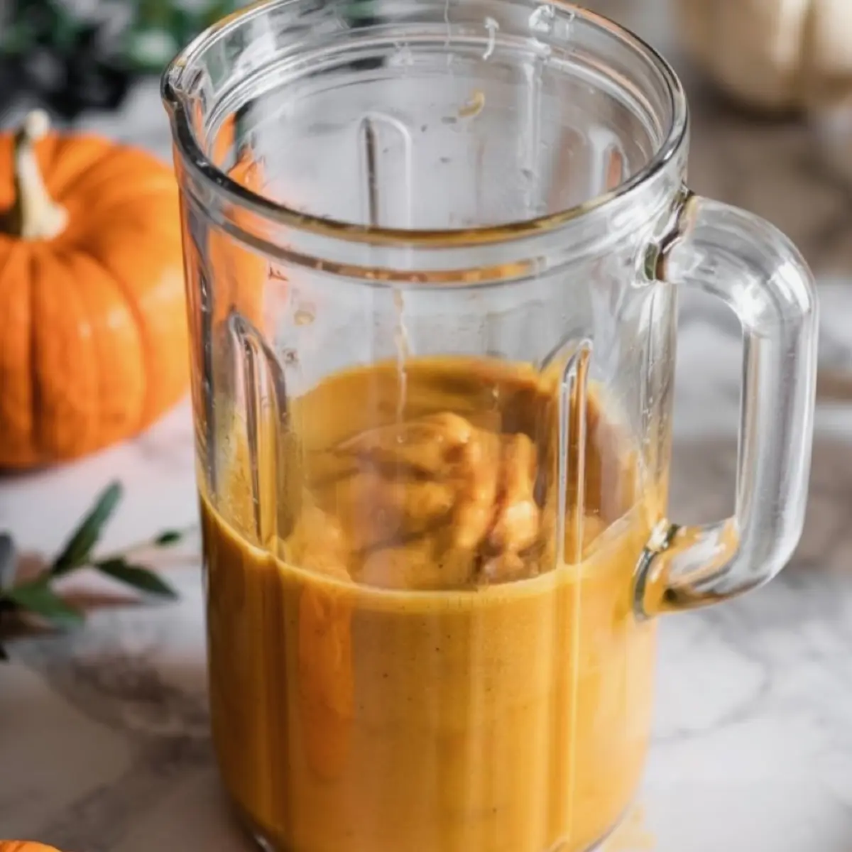 Blender jar filled with blended pumpkin smoothie mixture, showing a thick orange texture ready to be poured, with small pumpkins and leaves in the background.

