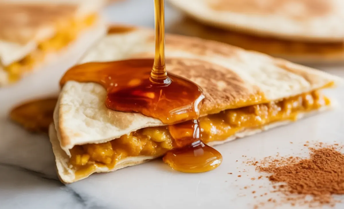 Close-up of a warm pumpkin spice breakfast quesadilla on a marble background, with thick maple syrup pouring over the folded tortilla filled with creamy pumpkin mixture.
