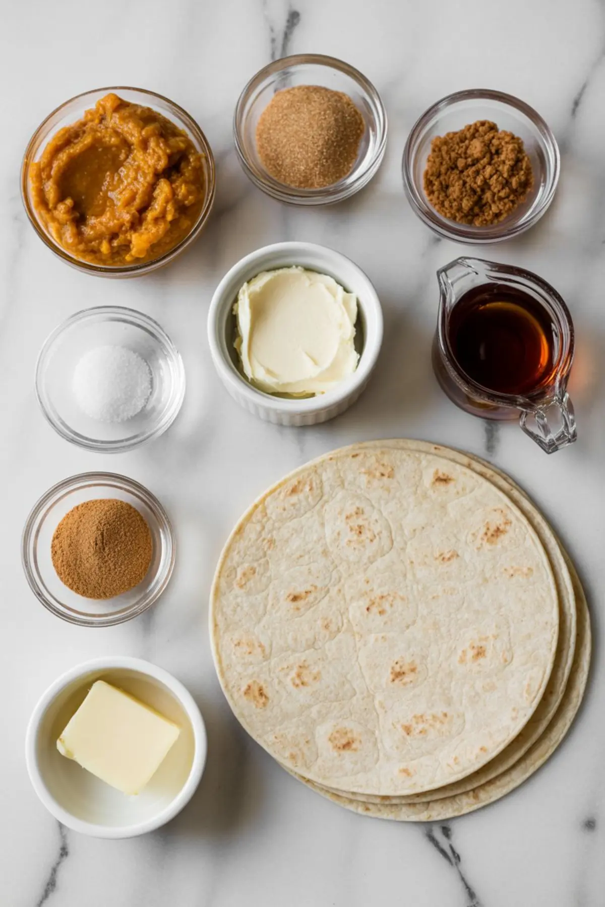 Flat lay of pumpkin spice breakfast quesadilla ingredients on a marble surface, including pumpkin purée, cream cheese, butter, flour tortillas, brown sugar, maple syrup, cinnamon, coconut sugar, and granulated sugar.
