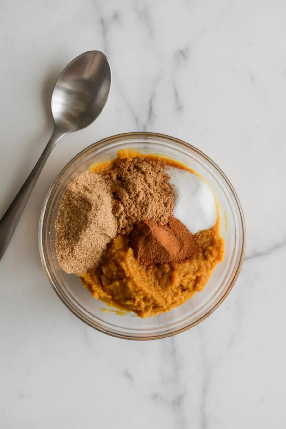 Bowl of pumpkin purée topped with cinnamon, brown sugar, coconut sugar, and granulated sugar on a white marble countertop, with a spoon placed beside it.
