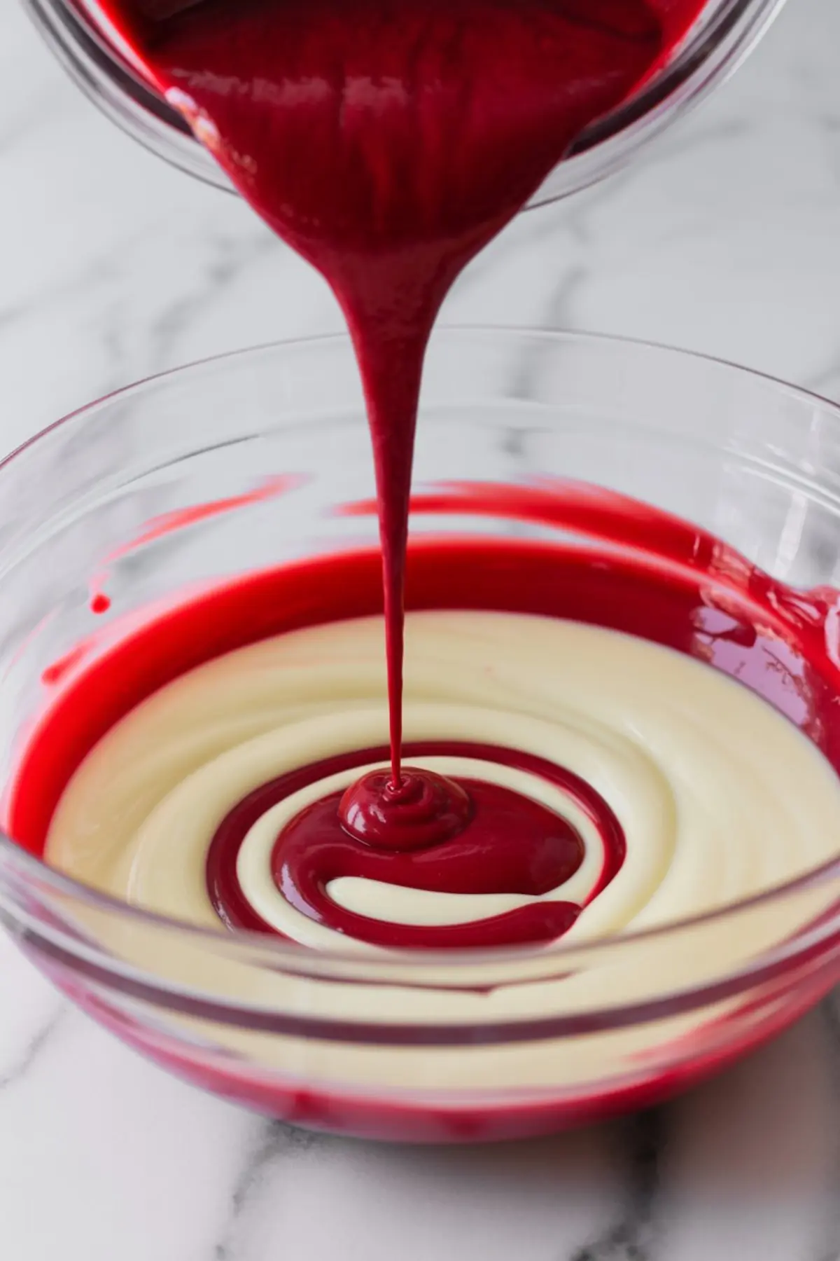 Raspberry puree being poured in a swirl pattern over white chocolate mixture in a glass bowl, creating a colorful marbled effect.