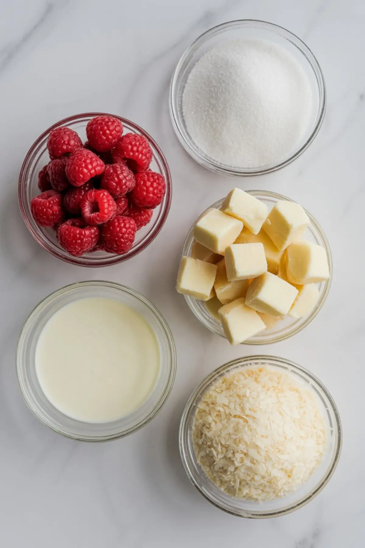 Flat lay of baking ingredients in glass bowls on a white marble surface, including fresh raspberries, granulated sugar, cubed butter, sweetened condensed milk, and shredded coconut.