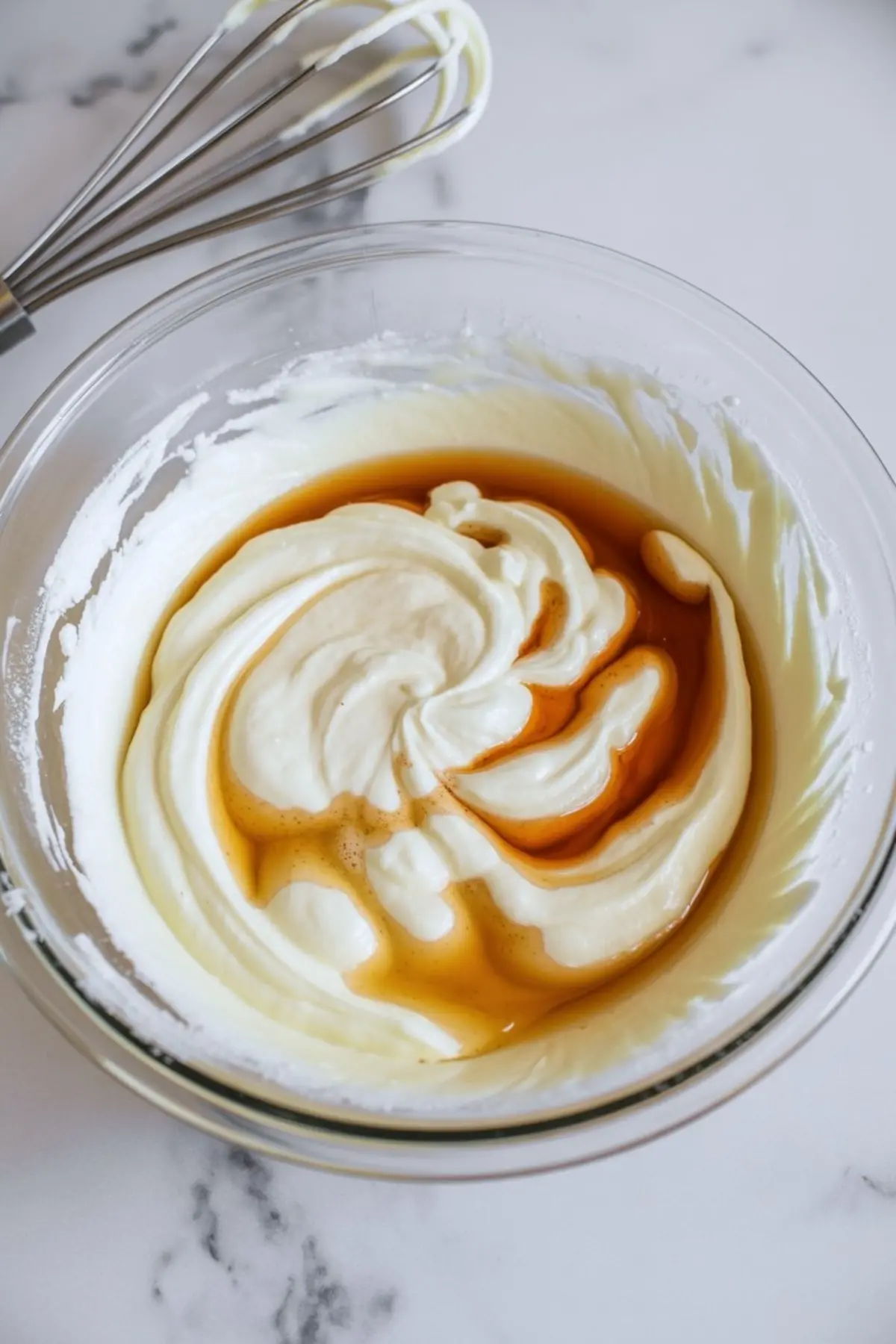 Whipped cream cheese and vanilla extract swirled together in a glass mixing bowl, placed on a marble countertop with a metal whisk beside it.